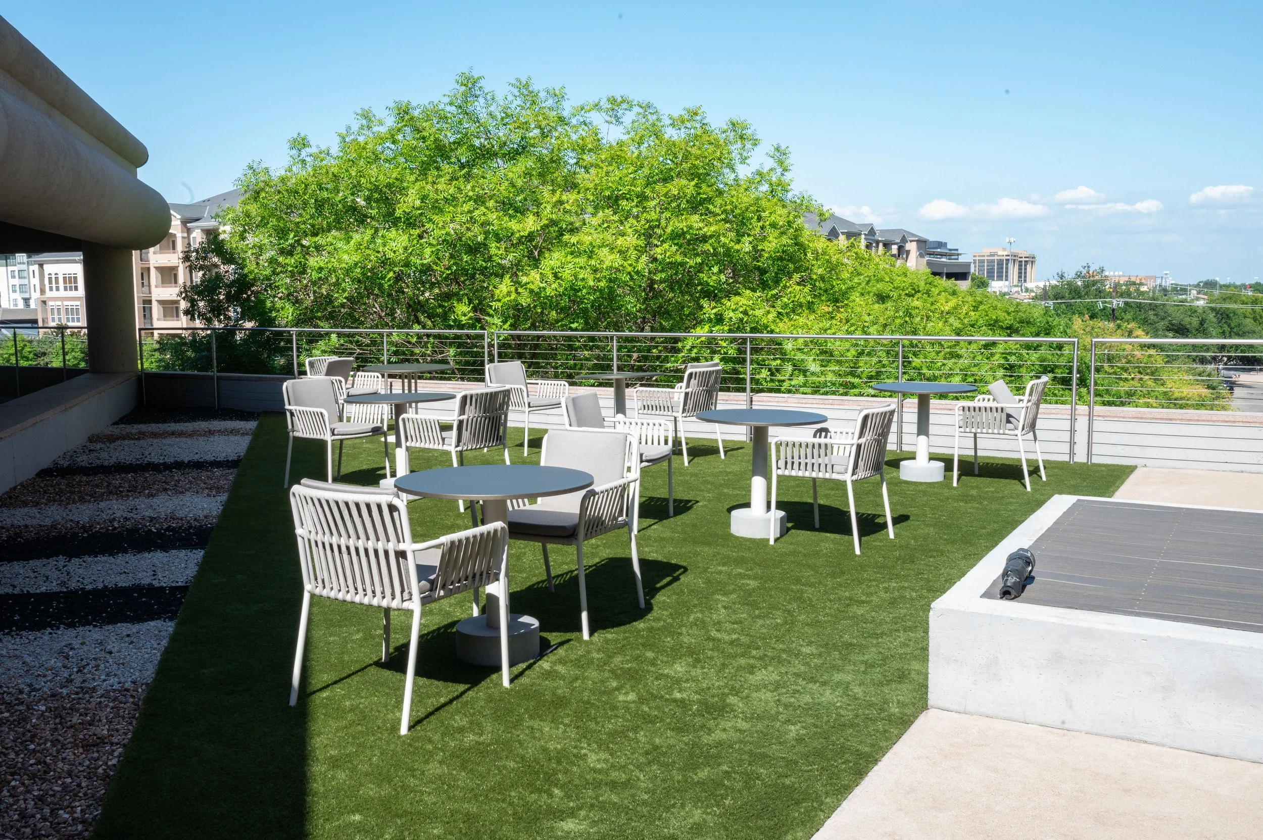 An outdoor rooftop patio with white tables and chairs, a green artificial grass surface, trees in the background, and a cityscape with buildings under a clear blue sky.