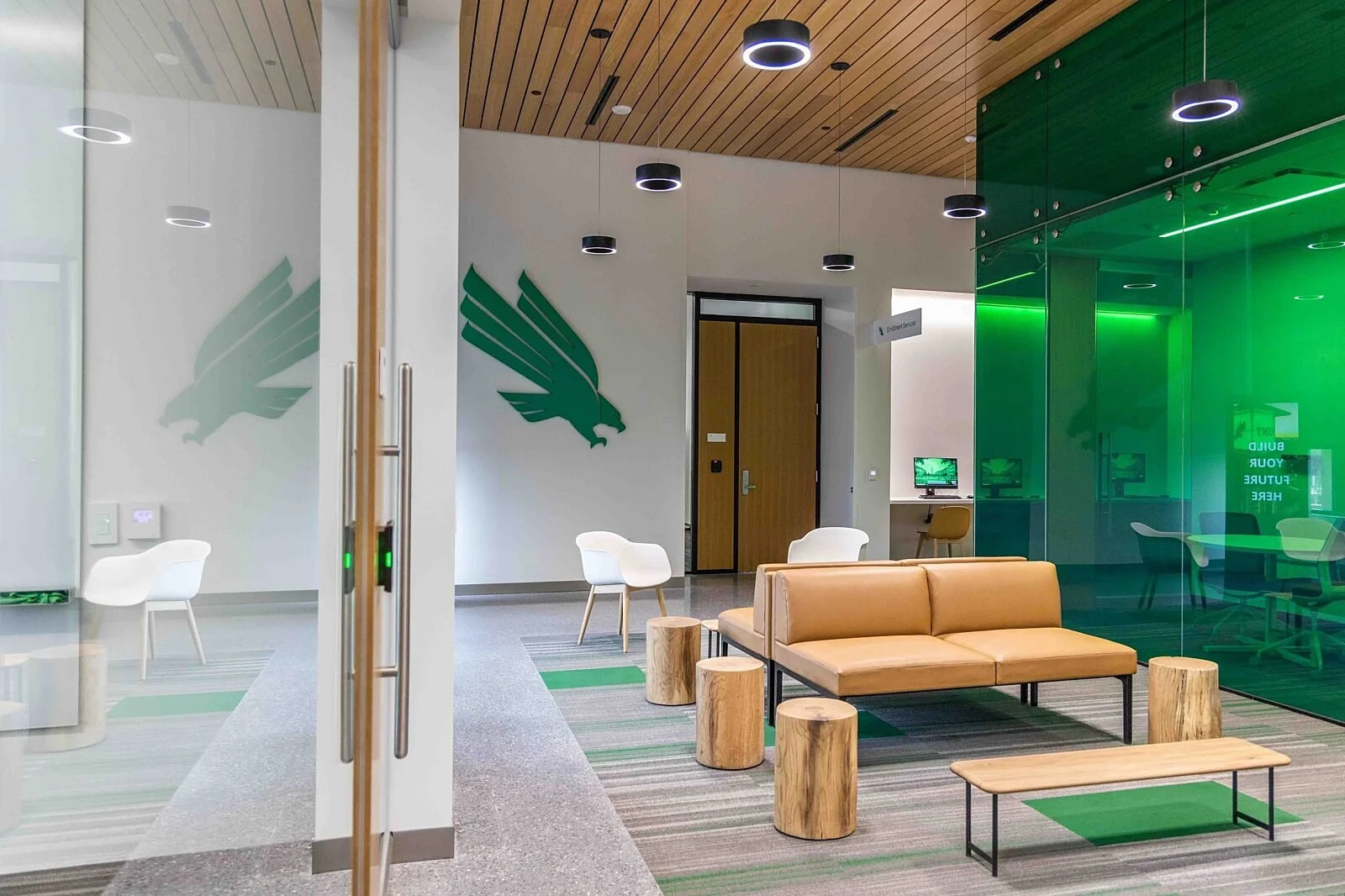 Modern office lobby with wooden ceiling, green glass wall, white and orange seating, and eagle decals on white walls.
