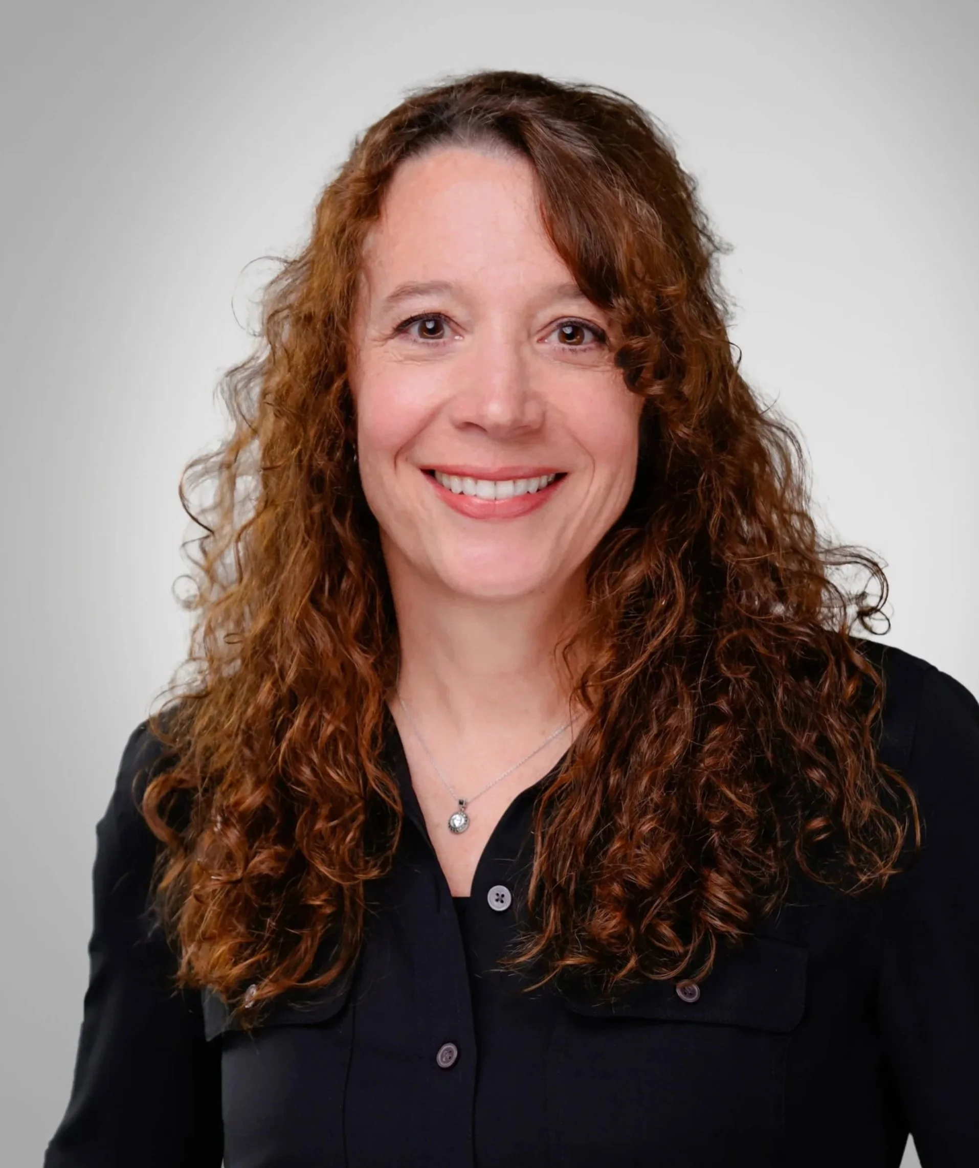 A woman with curly red hair smiling, wearing a black blouse with buttons and a silver necklace with a round pendant, against a plain light gray background.