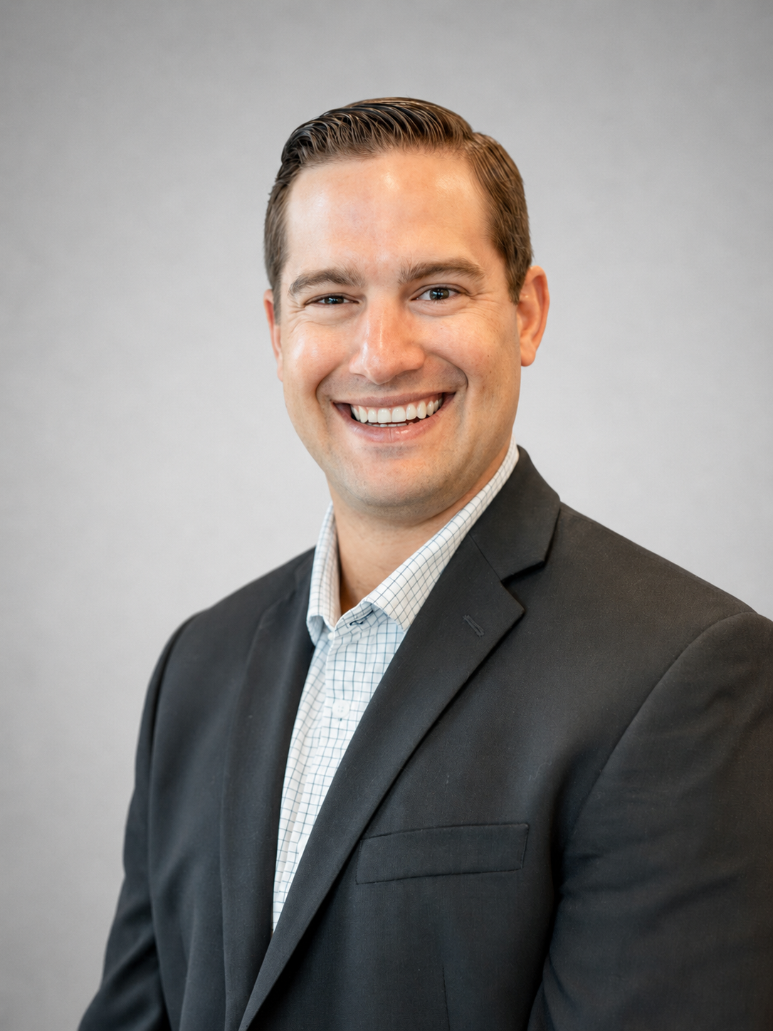 Headshot of a smiling man wearing a dark suit and a light checkered shirt against a gray background.