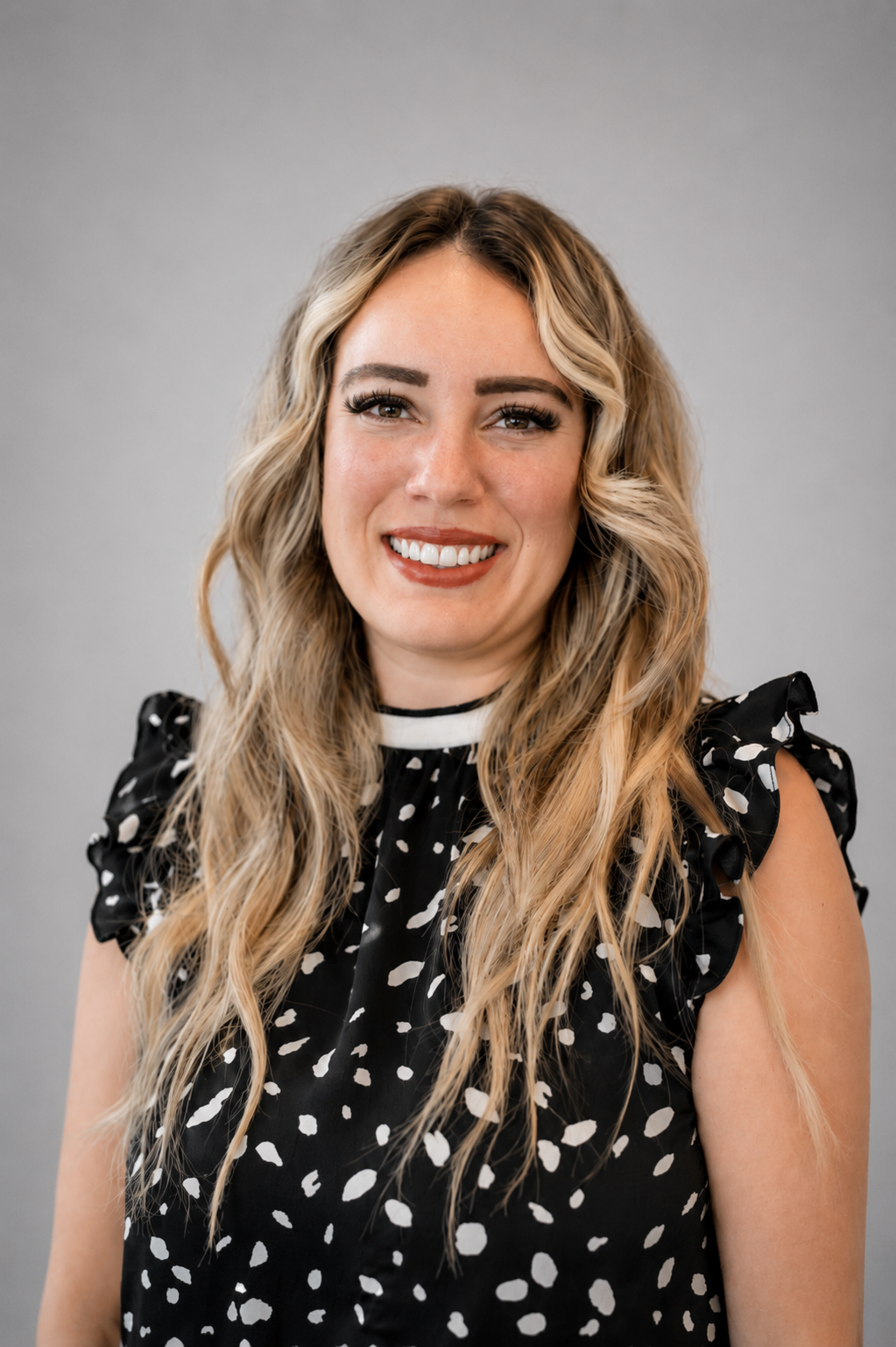 A woman with long, wavy blonde hair, smiling, wearing a black dress with white polka dots and ruffled sleeves, posing in front of a plain gray background.