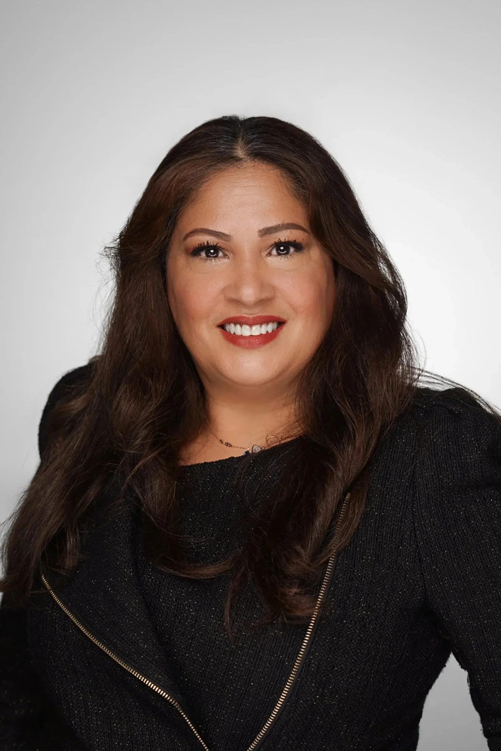 A woman with long brown hair wearing a black top and smiling, against a plain light background.