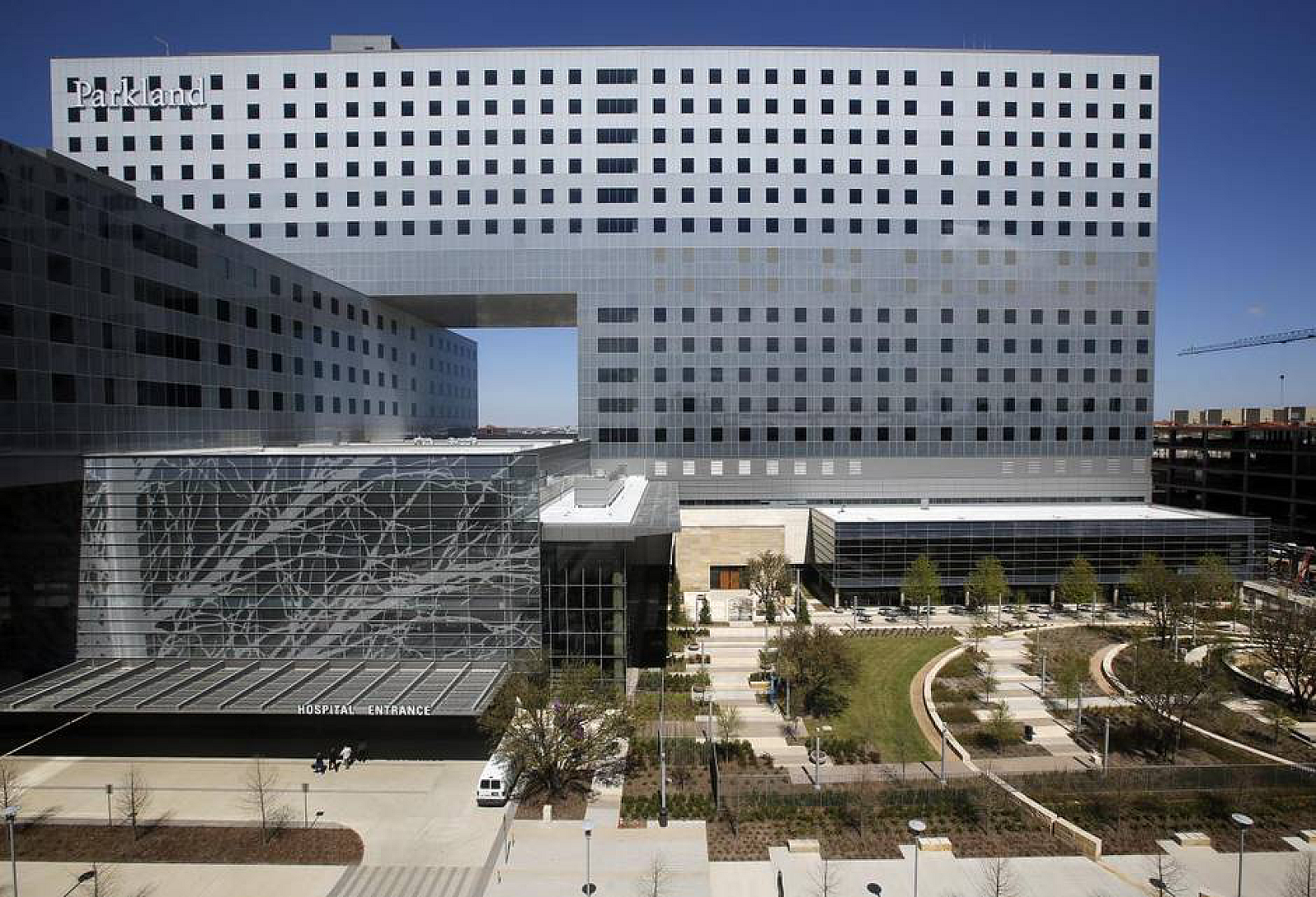 Modern hospital building with a large, rectangular main structure and glass entrance area, surrounded by a landscaped outdoor space with trees and pathways.