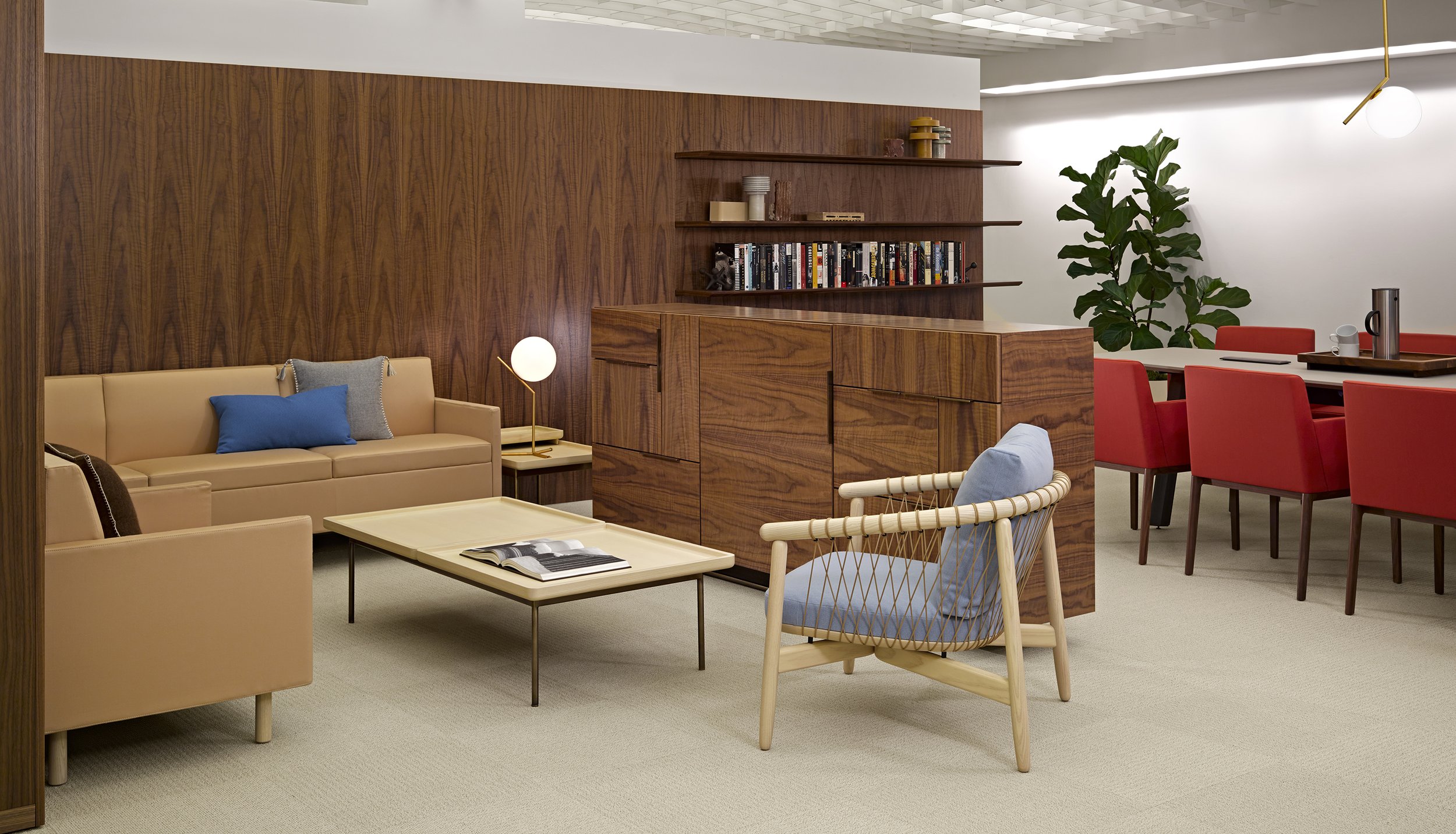 Modern living room with beige sofa, blue and gray pillows, wooden cabinets, a white coffee table with an open magazine, another armchair with a light blue cushion, and a large green houseplant. In the background, a dining area with a white table, red chairs, and a wooden tray with a coffee pot and mugs. Wooden and white shelves with decorative objects and books are on the wall.