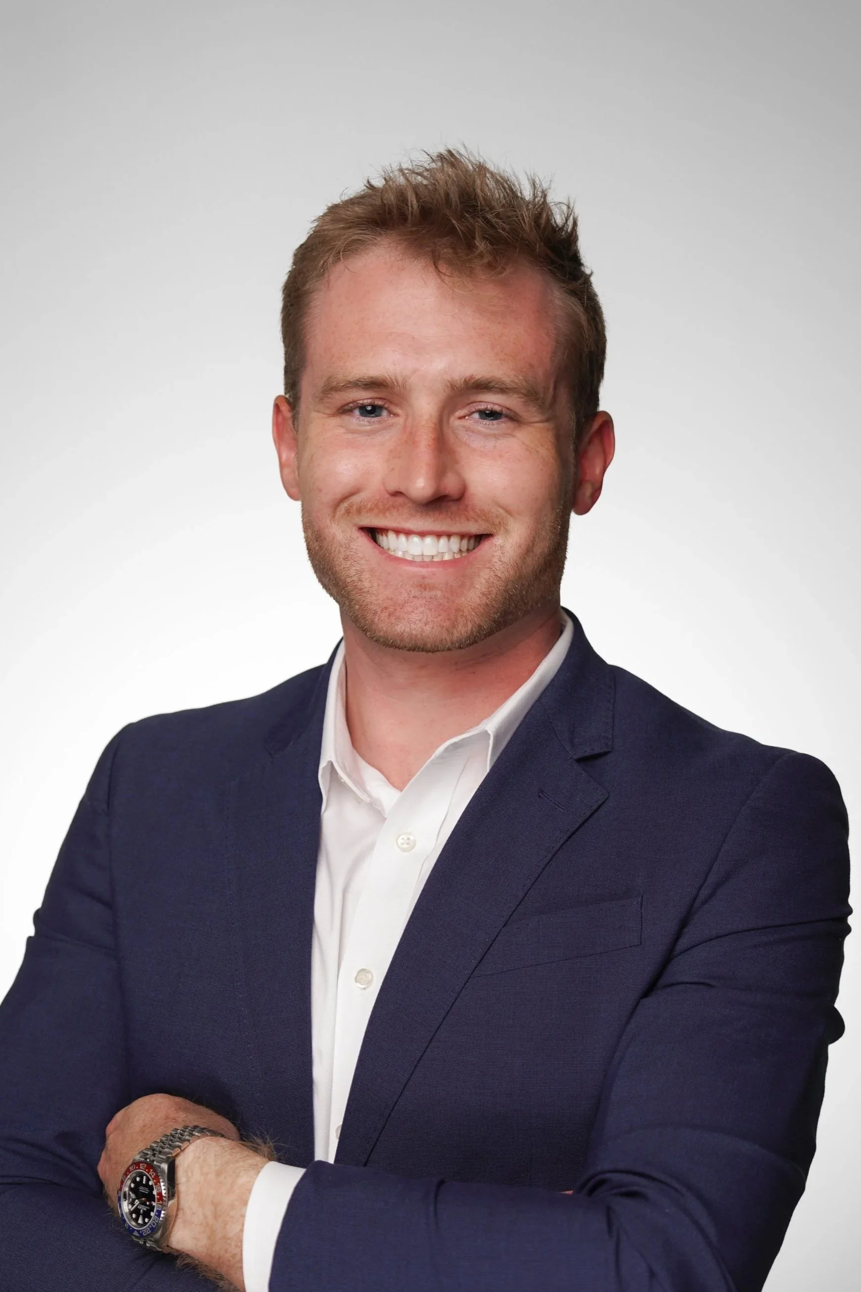 A young man in a navy blue suit and white shirt smiling with arms crossed, wearing a silver watch, against a plain gray background.
