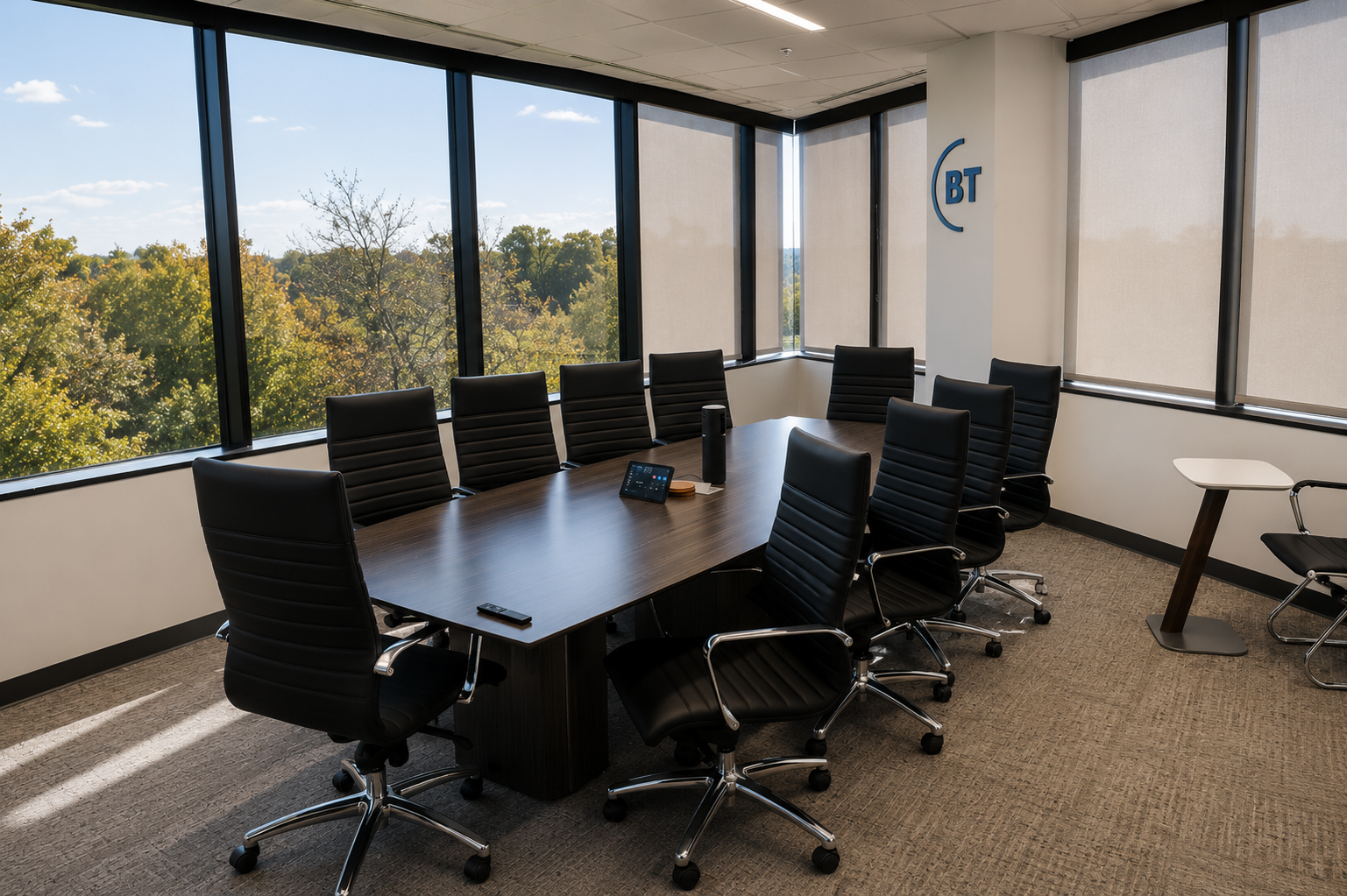 Empty modern conference room with a large wooden table, black leather chairs, and large windows with beige shades, overlooking trees and a blue sky.