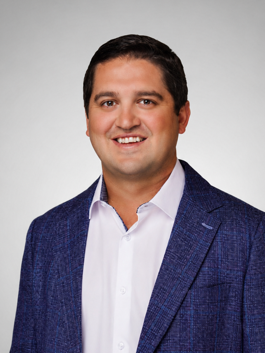 Portrait of a man with dark hair wearing a white shirt and a dark blue blazer, smiling against a light gray background.