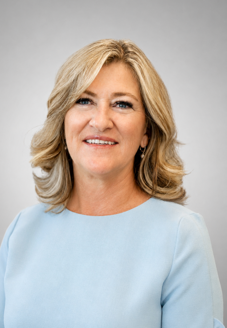 A middle-aged woman with blonde, curled hair and blue eyes, smiling gently, wearing a light blue top and pearl earrings against a plain light gray background.