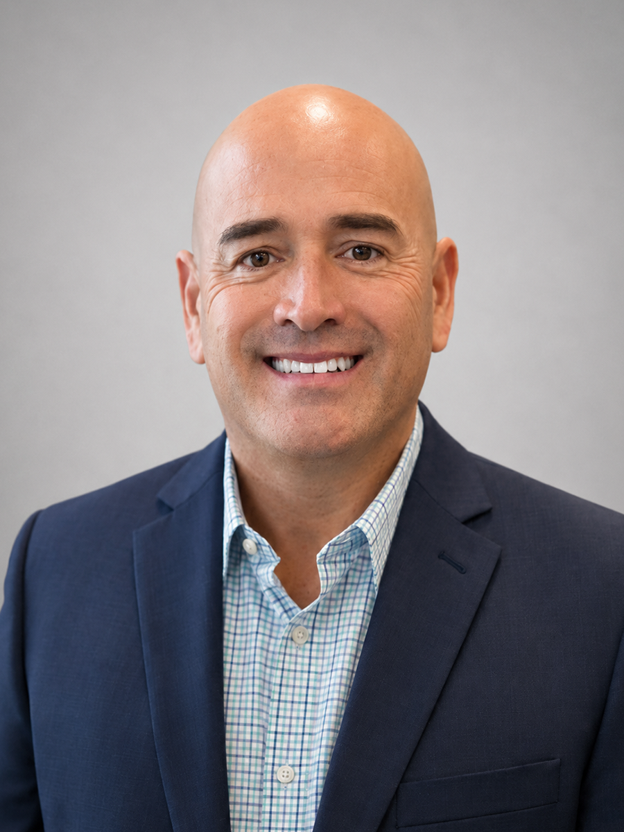 A smiling man with a shaved head wearing a dark suit jacket and a patterned dress shirt against a grey background.