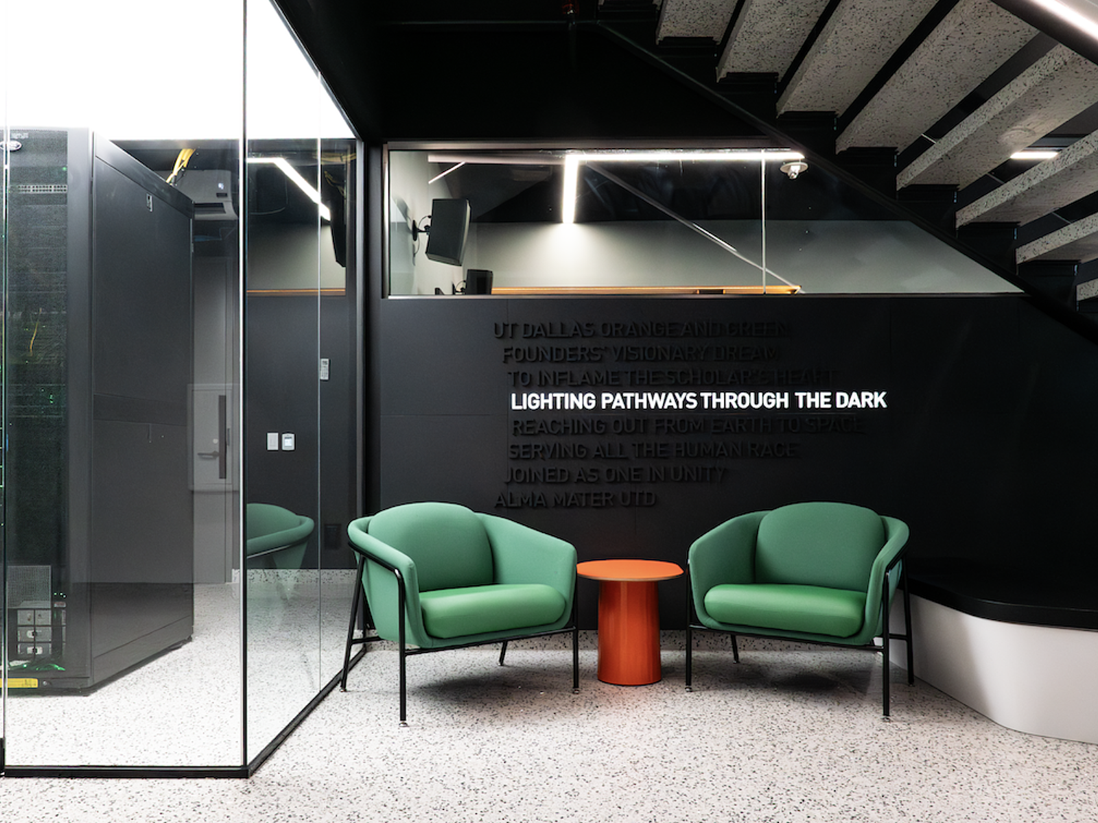 Interior of a modern building with two green armchairs separated by a small orange table, black wall with text about lighting pathways through the dark, and a staircase with exposed concrete steps.