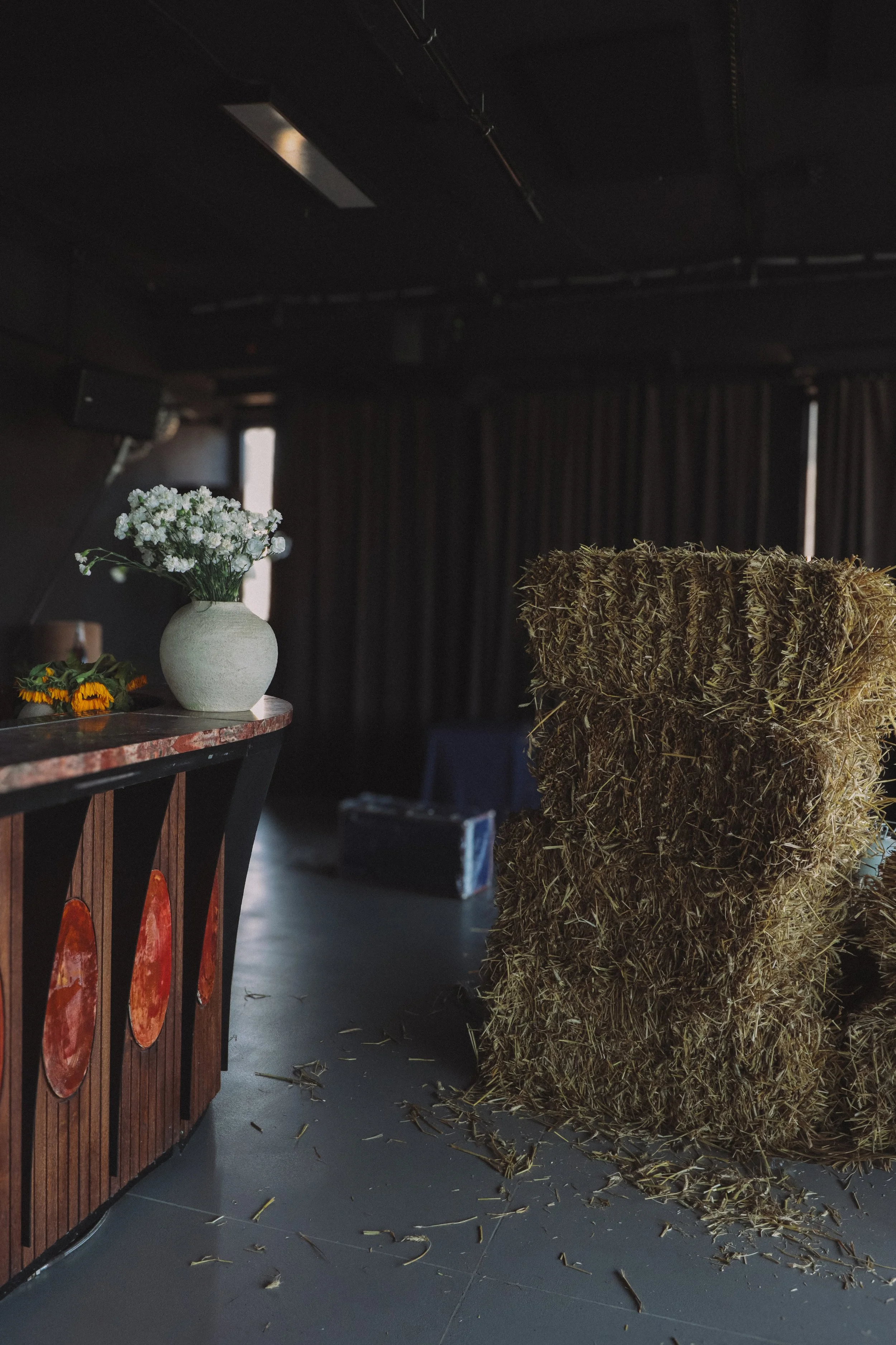 Interior of a space with a bar to the left featuring a decorative pizza design, a vase of white flowers on the bar, a hay bale to the right, and scattered hay on the floor. Dark curtains cover the back wall, and a window lets in natural light.