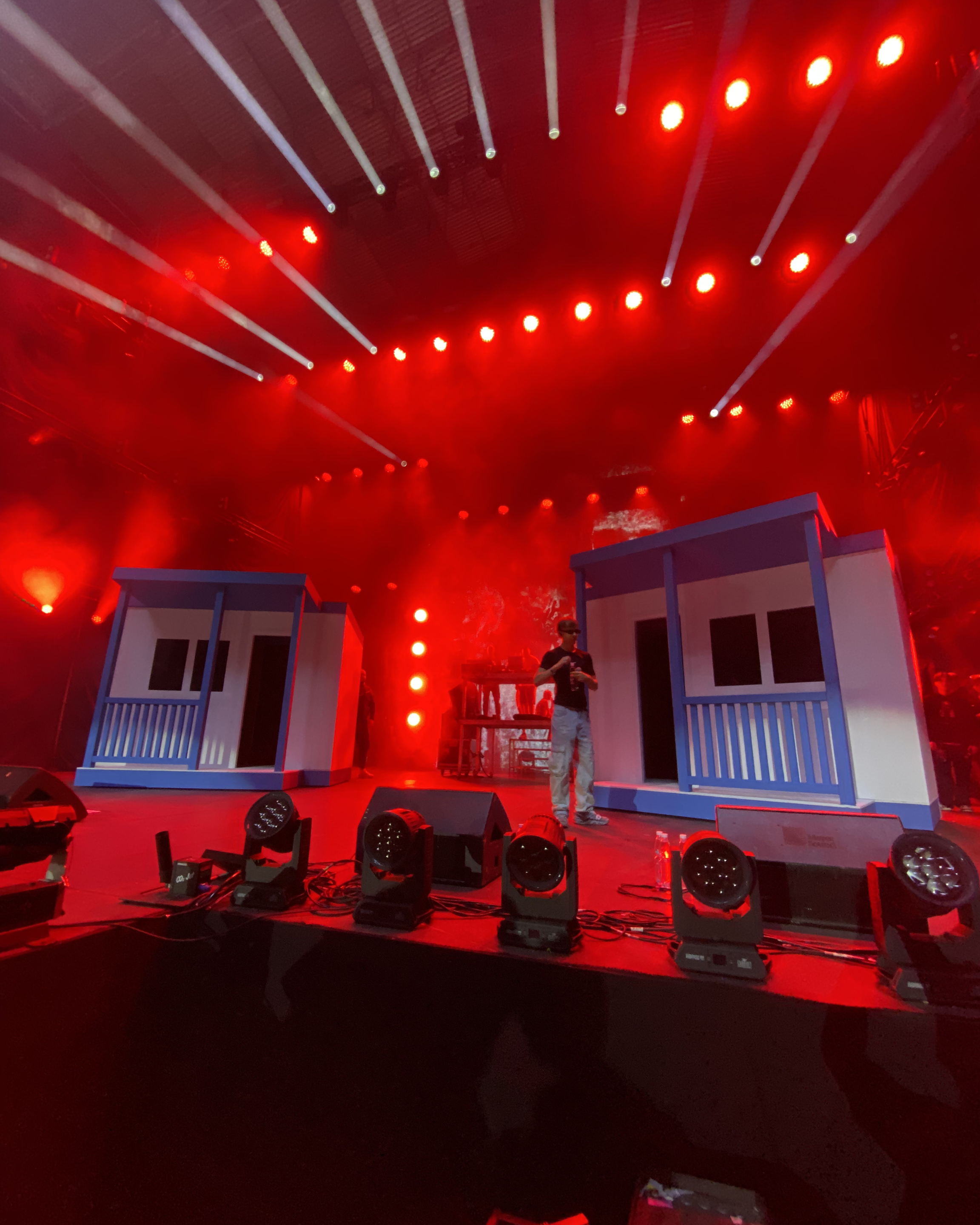 Stage with set pieces resembling houses, colorful lighting, and a performer in dark clothing with a visor or sunglasses.