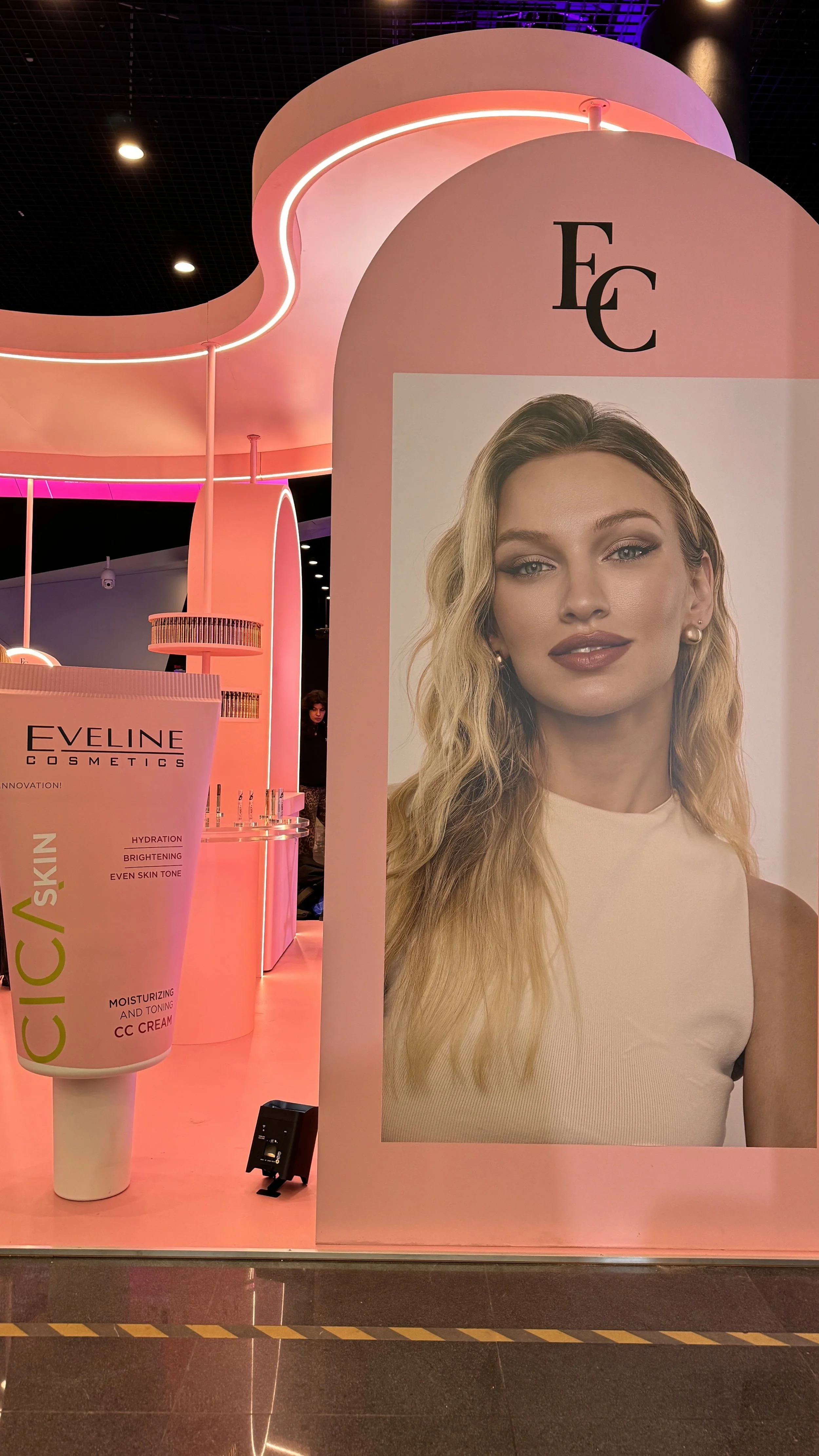 A large advertisement featuring a woman's face with blonde hair and natural makeup, promoting Eveline Cosmetics. The display is illuminated with pink and purple lighting, with parts of the cosmetic products visible nearby.