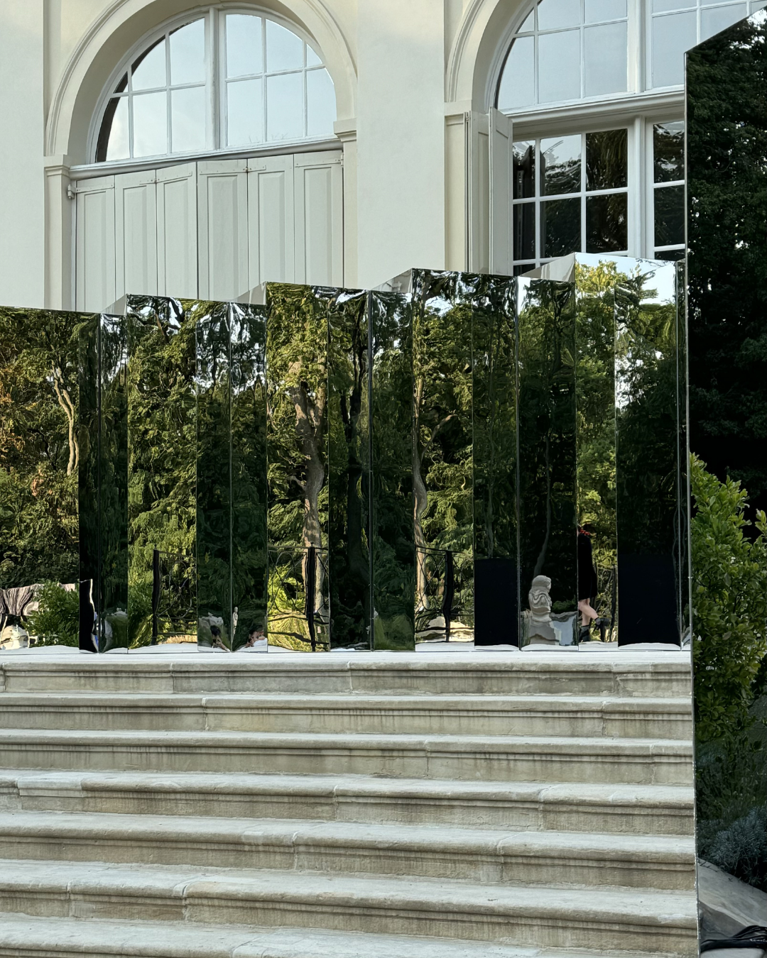 Mirrored installation on steps in front of a large white building with arched windows and white shutters.