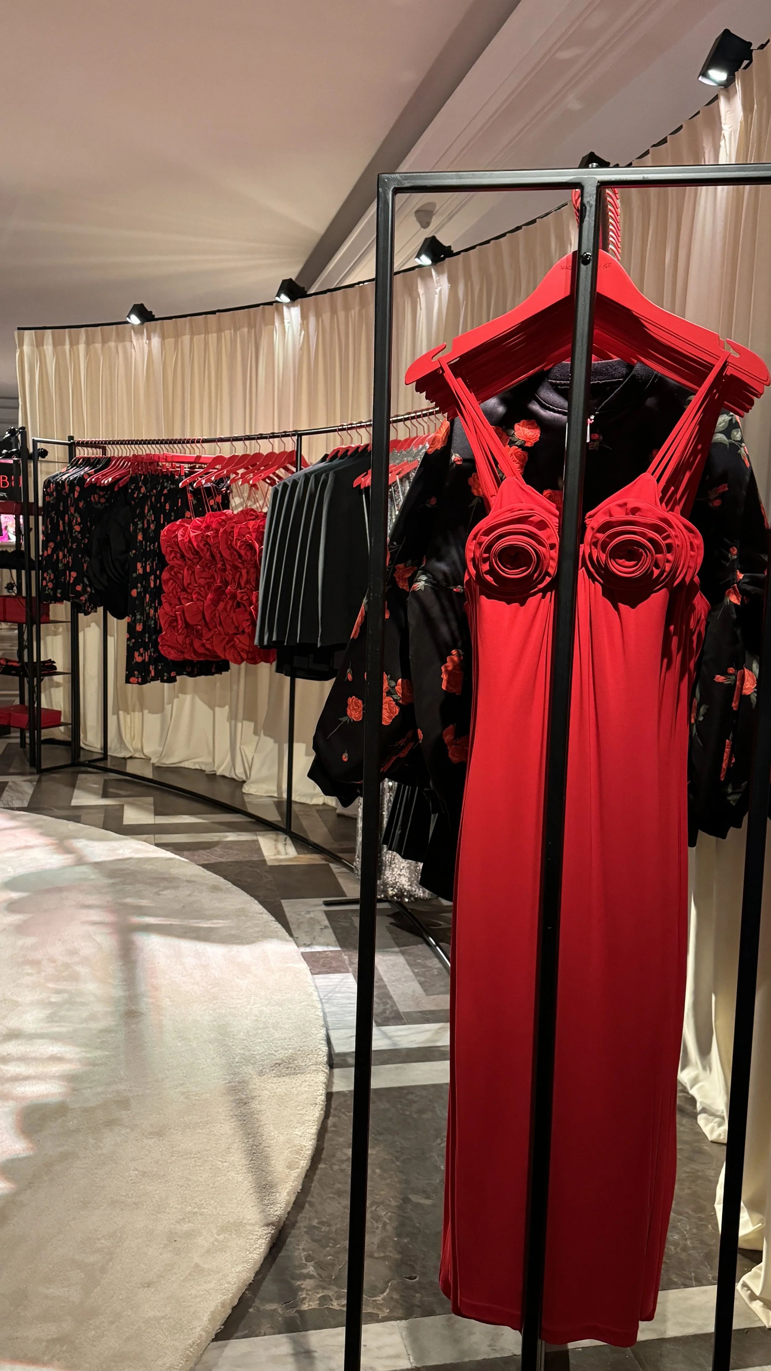 Red dress displayed on a hanger in a clothing store with various black and floral garments on racks in the background.
