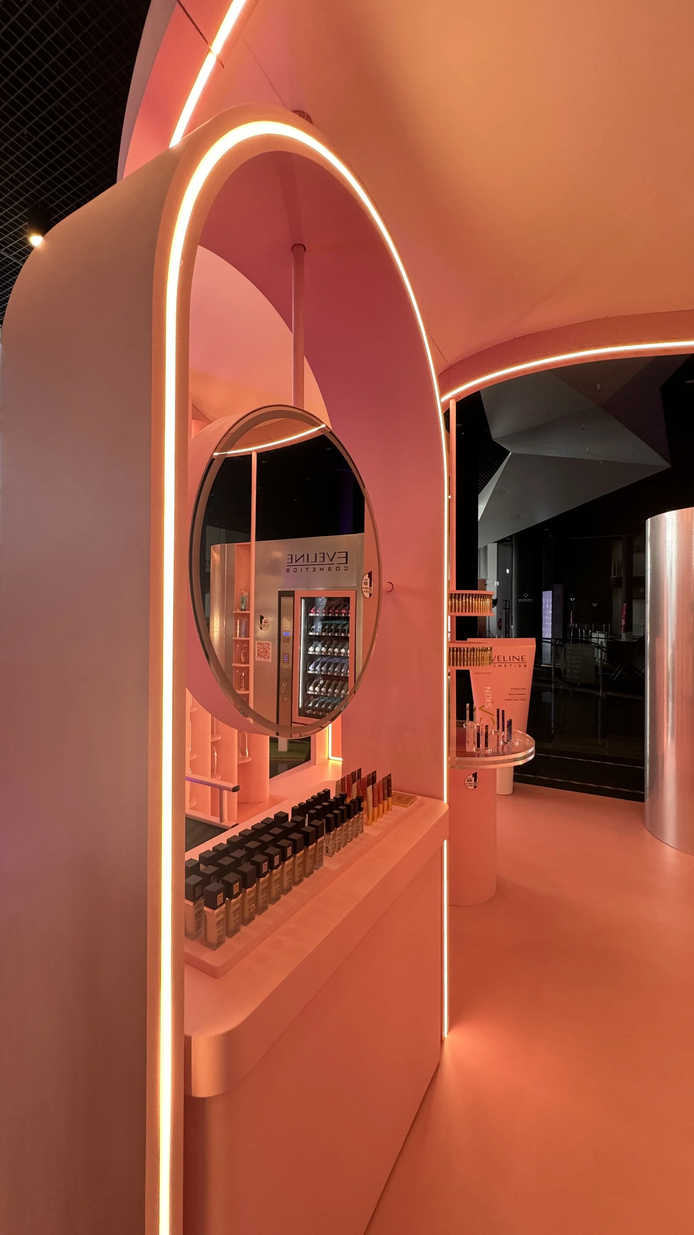 Interior of a cosmetics store with pink lighting and display shelves holding makeup products, including foundation bottles, with a curved modern design.