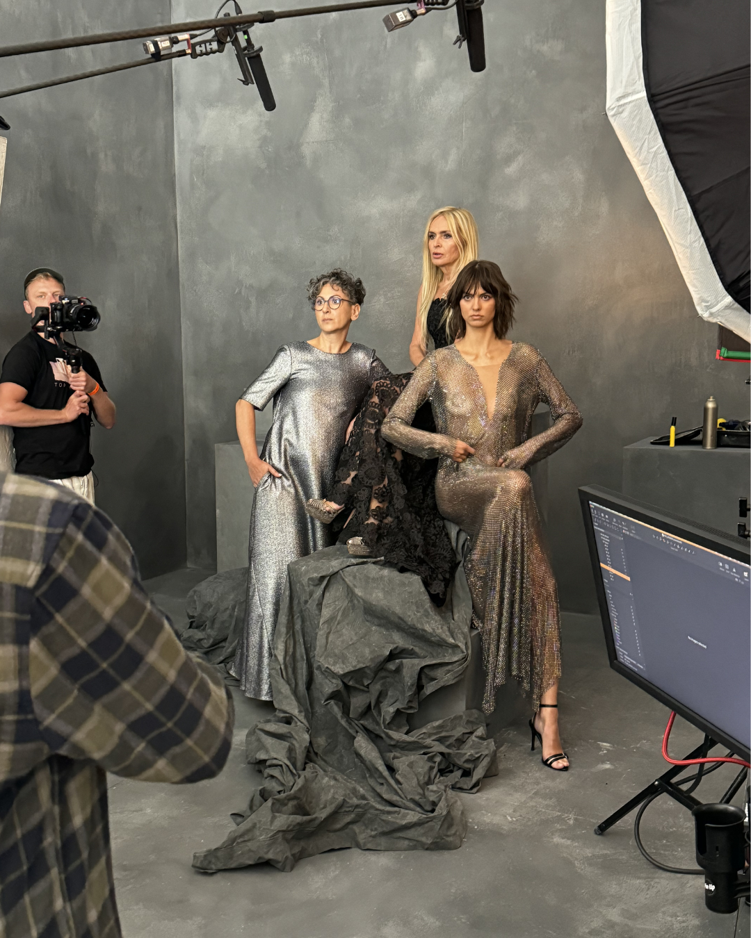 Four women in metallic and black dresses posing for a photoshoot in a studio with gray walls and equipment around.