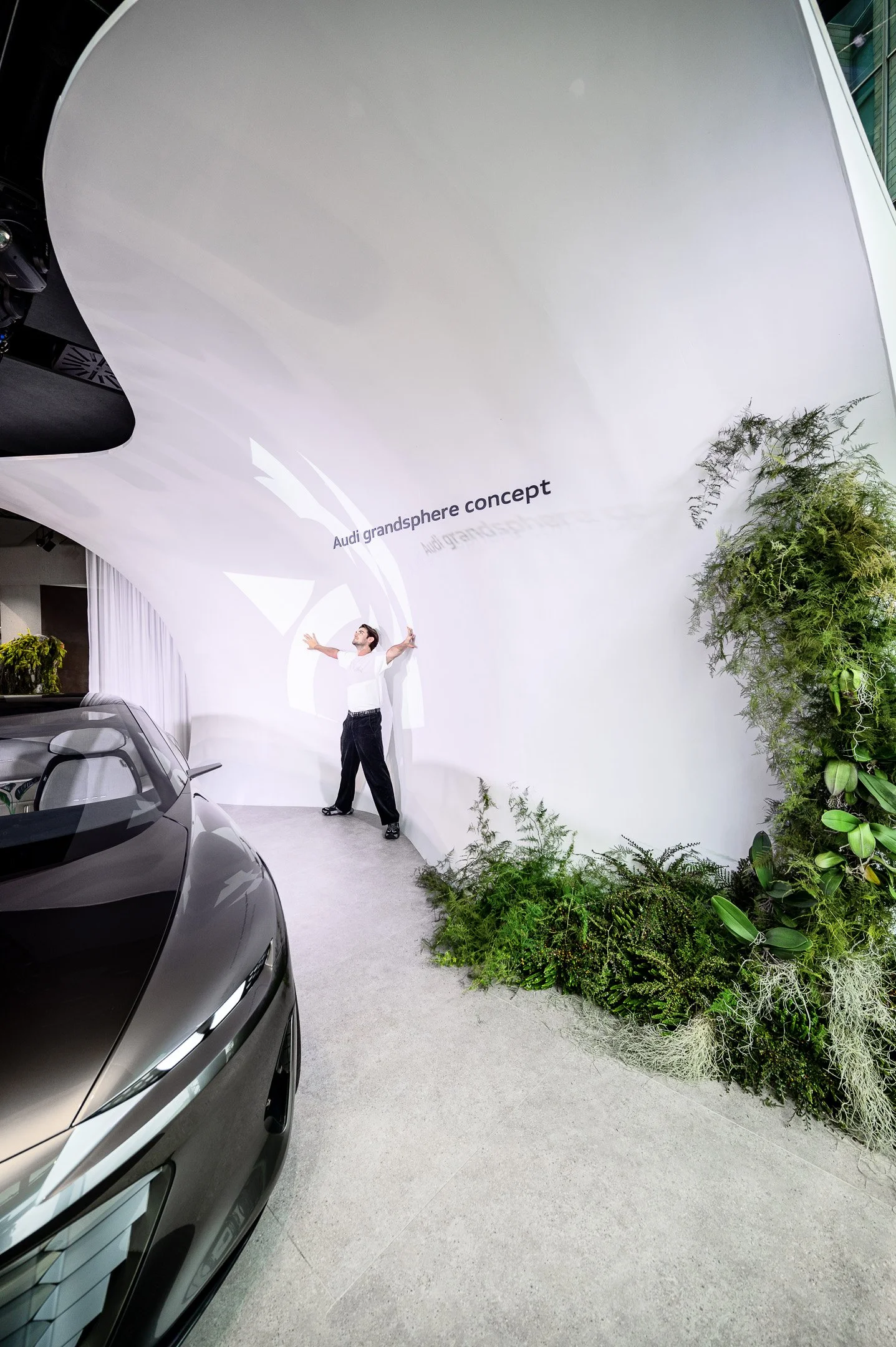 A man standing in front of a white backdrop with the words 'Audi grandsphere concept' written on it. There are cars in the foreground and green plants on the right side of the backdrop.
