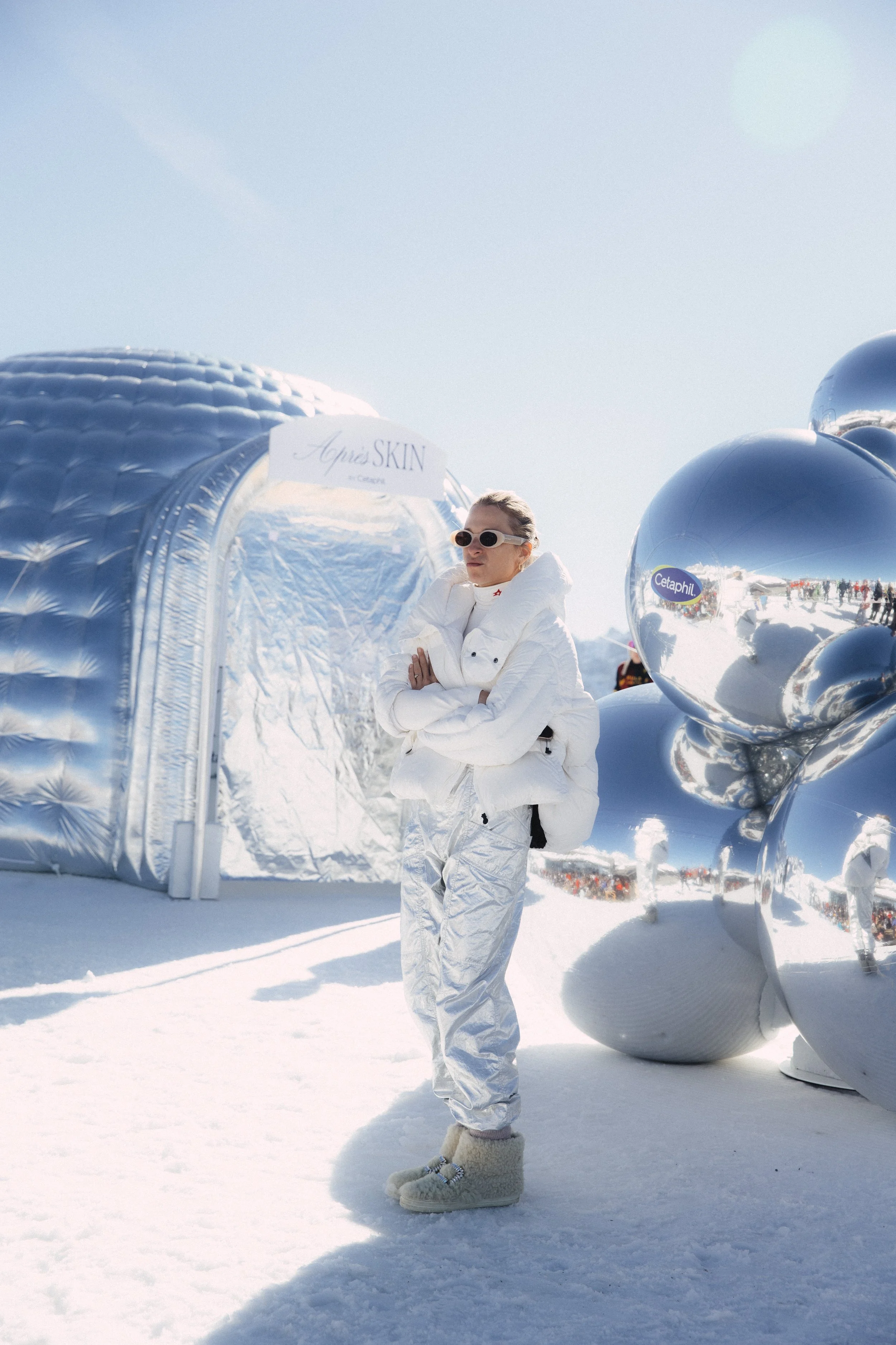 A woman wearing white sunglasses, a white puffy jacket, and shiny silver snow boots standing on snow with reflective balloons nearby and a metallic igloo with a sign that reads 'Apiis SKIN' in the background.