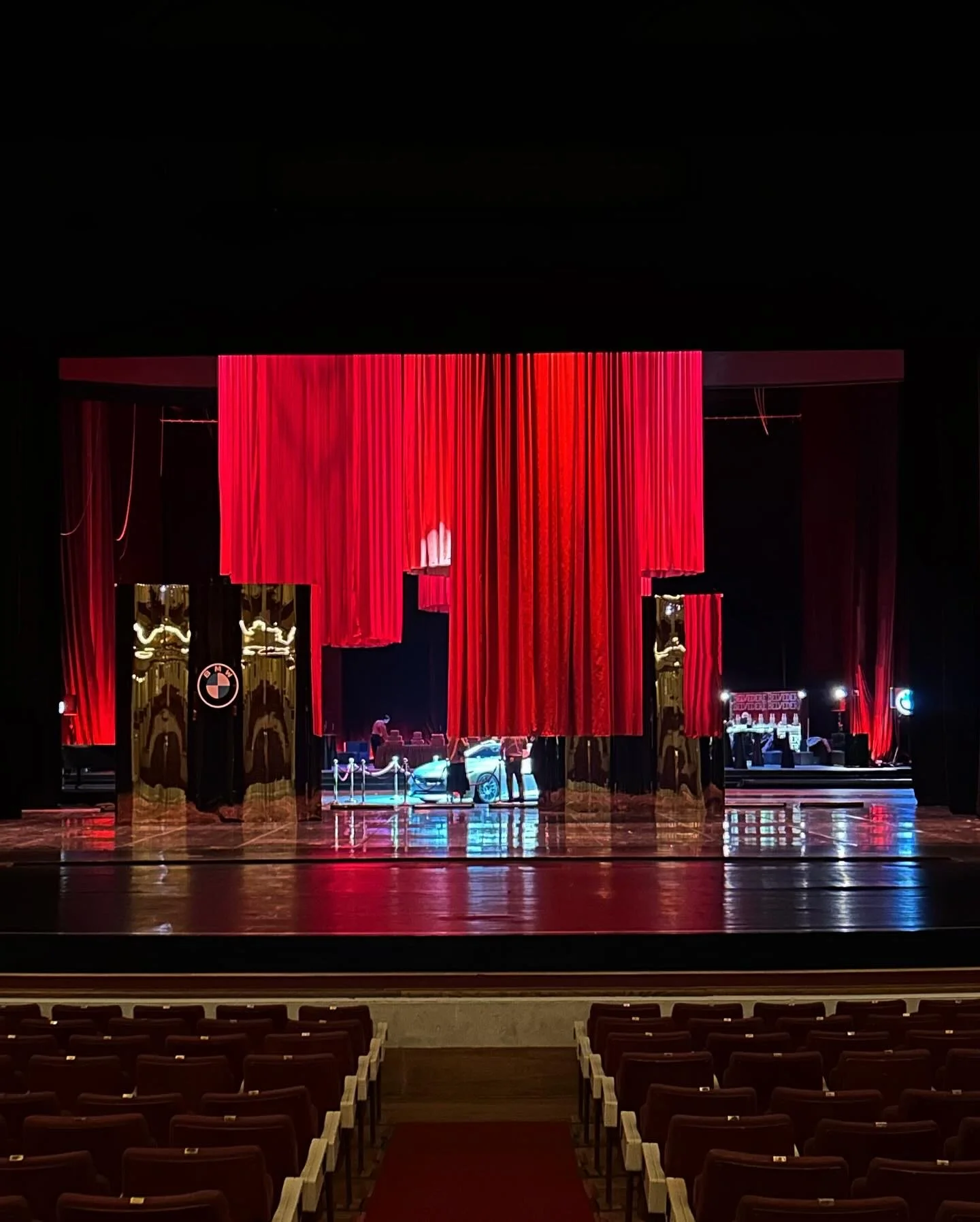 An empty theater with red curtains, a stage with a luxury car, and maroon seats in the foreground.