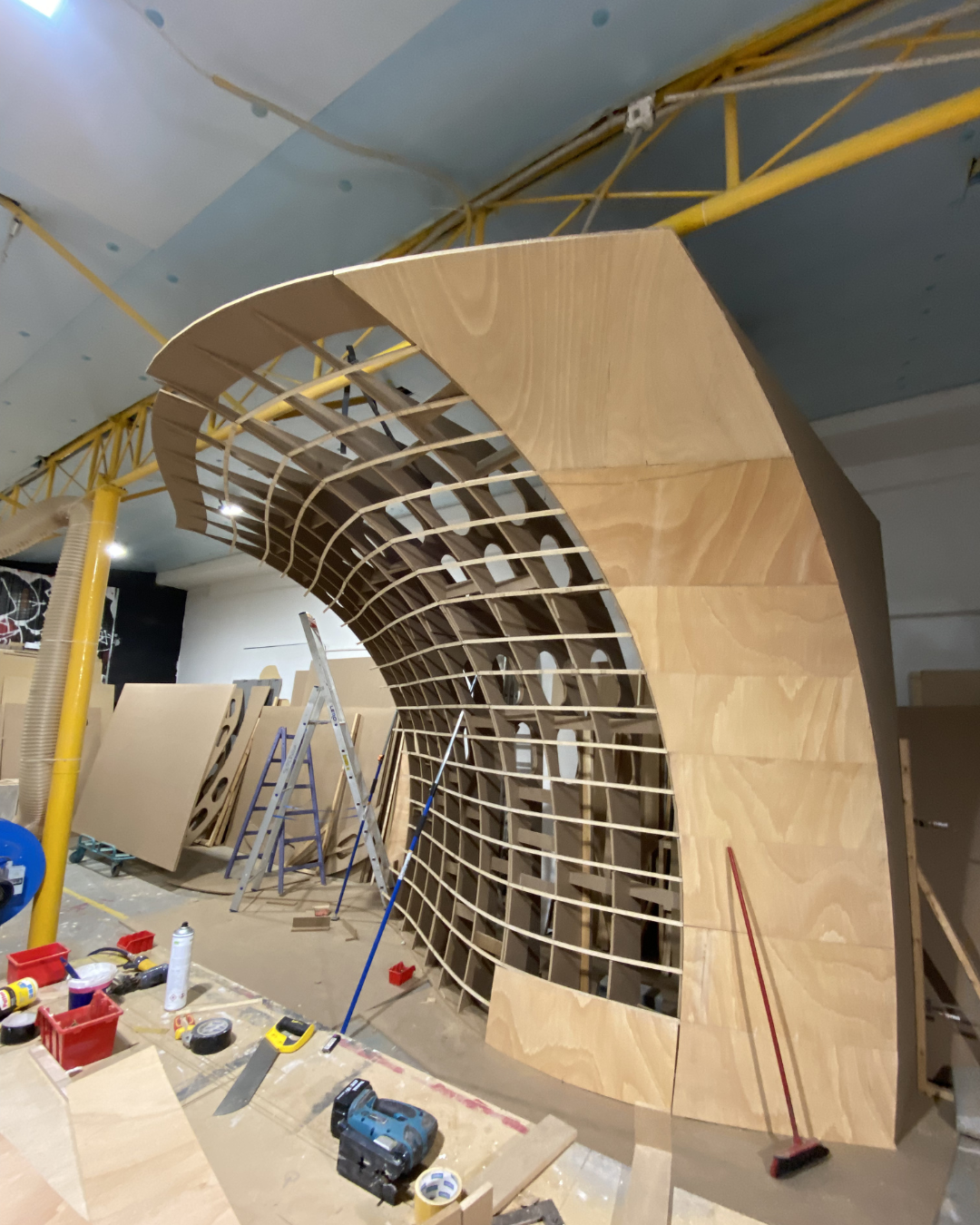 Wooden structure with curved framework in a workshop, surrounded by tools, ladders, and construction materials.