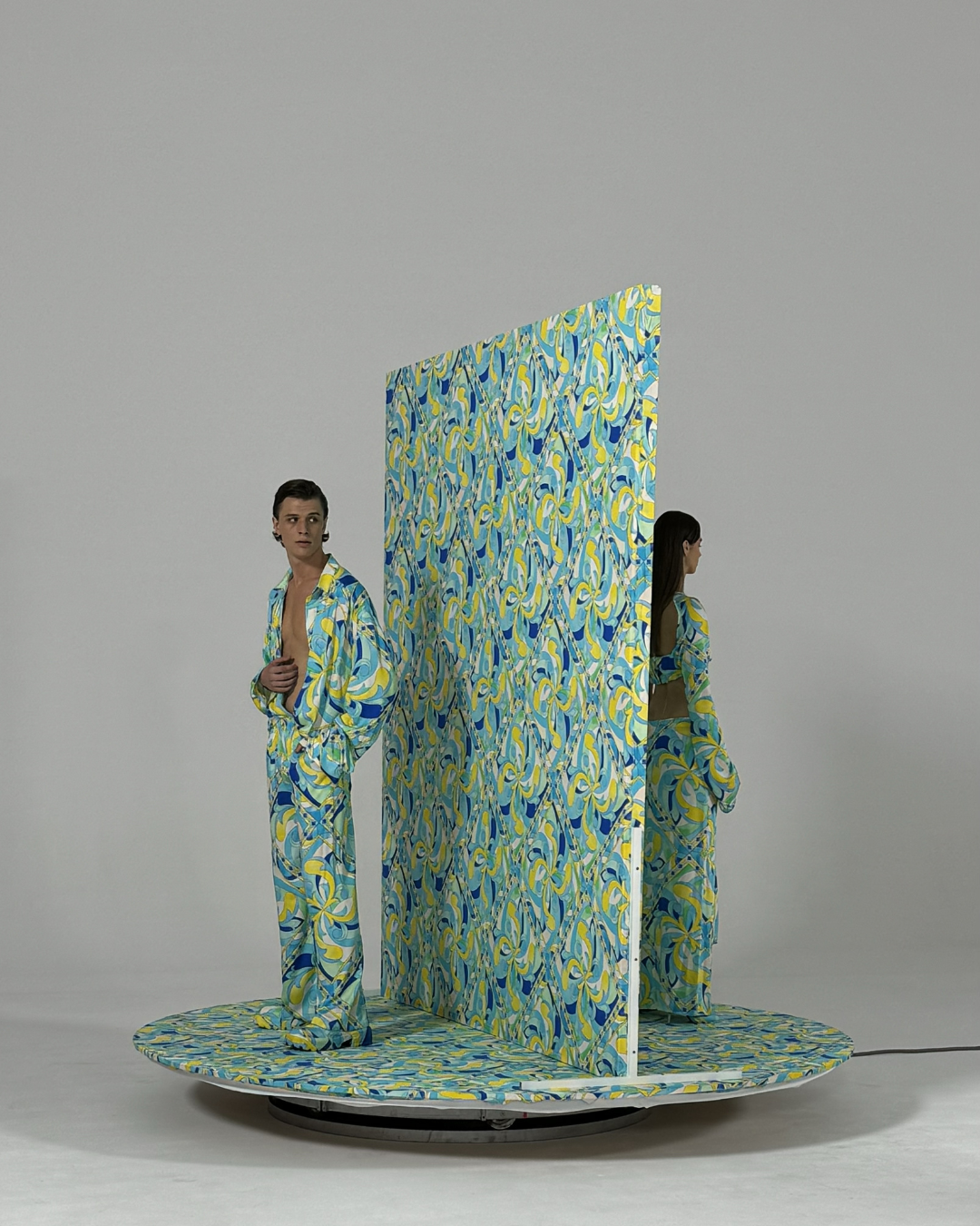 Two people in patterned clothing are positioned on a rotating platform with a large, matching patterned partitioning screen between them. One person is facing forward and the other is shown from the side.
