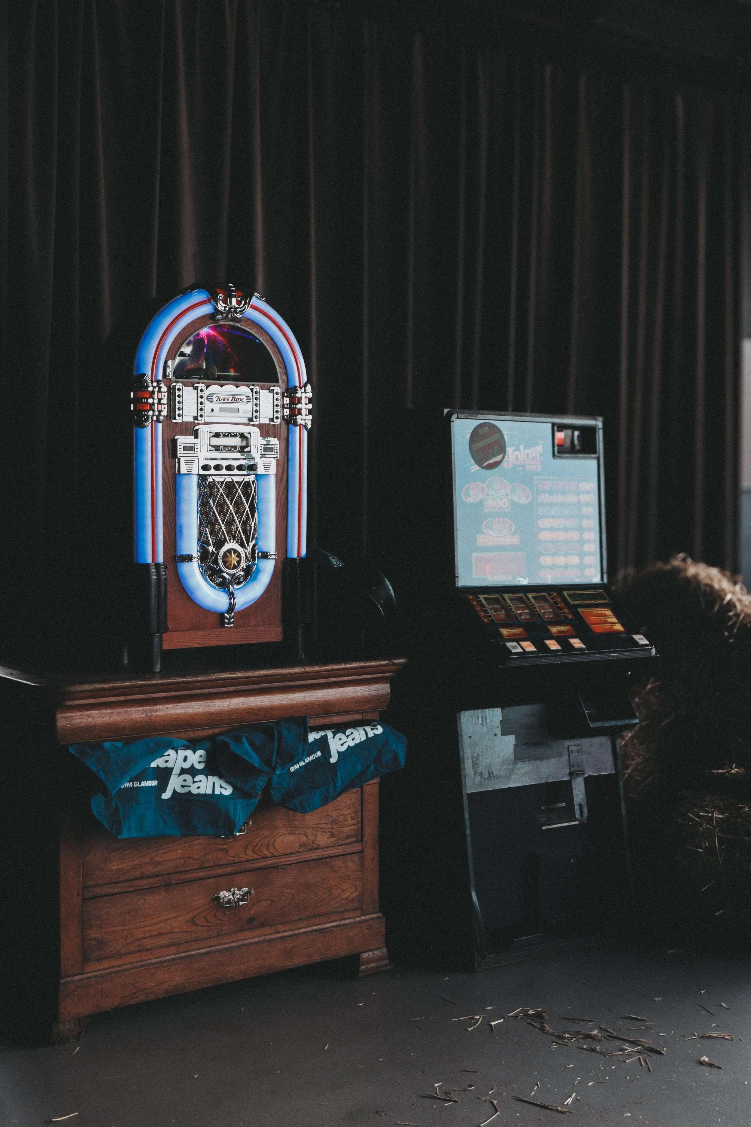 A vintage jukebox on a wooden cabinet with a black curtain background, nearby a digital jukebox screen, and a pair of blue jeans hanging from the cabinet drawer handle, with scattered debris on the floor.