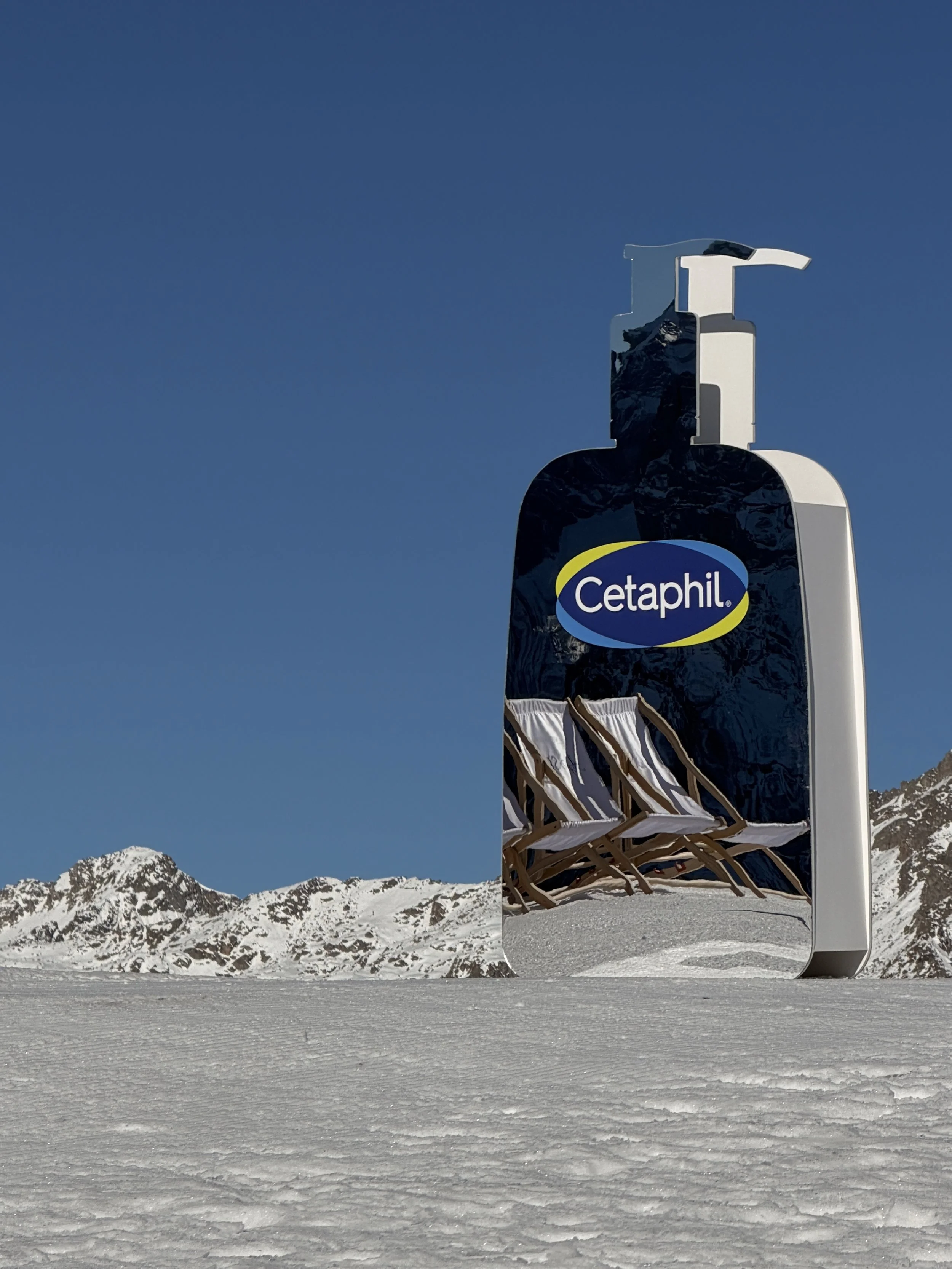 A large reflective sign shaped like a bottle of Cetaphil lotion stands on snow in a mountainous landscape with snow-covered peaks in the background. The sign's surface mirrors the snow and mountains.