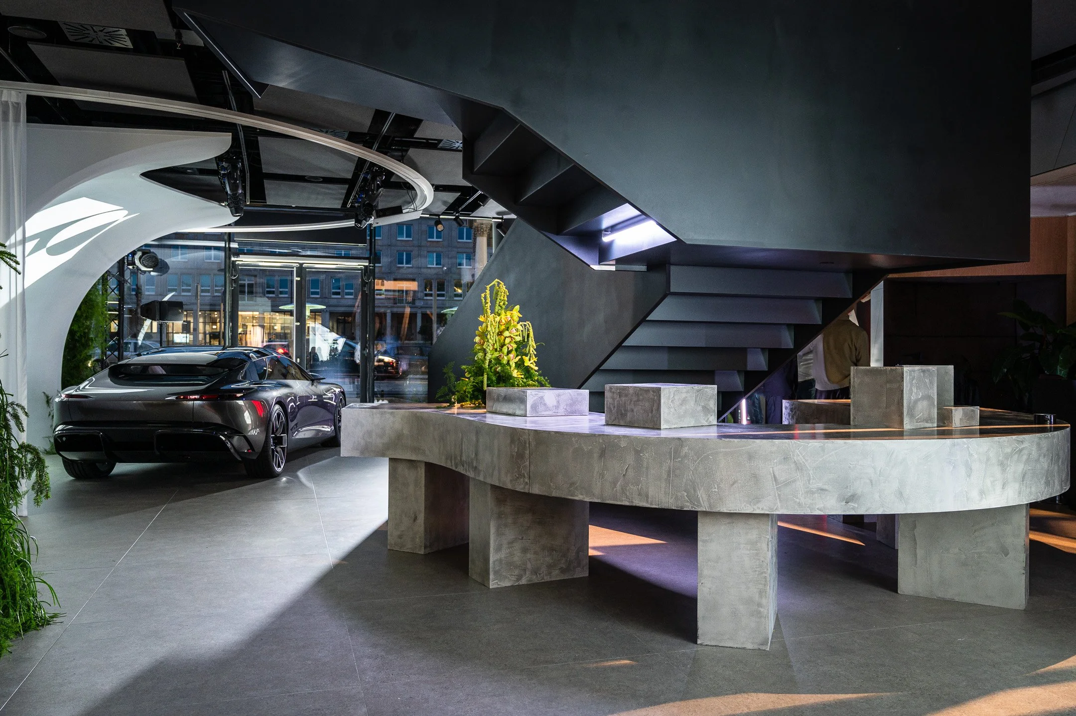 A modern indoor showroom with a dark interior, featuring a silver luxury car parked near large glass windows, a concrete reception desk, a staircase with black steps, and some green plants and flowers.