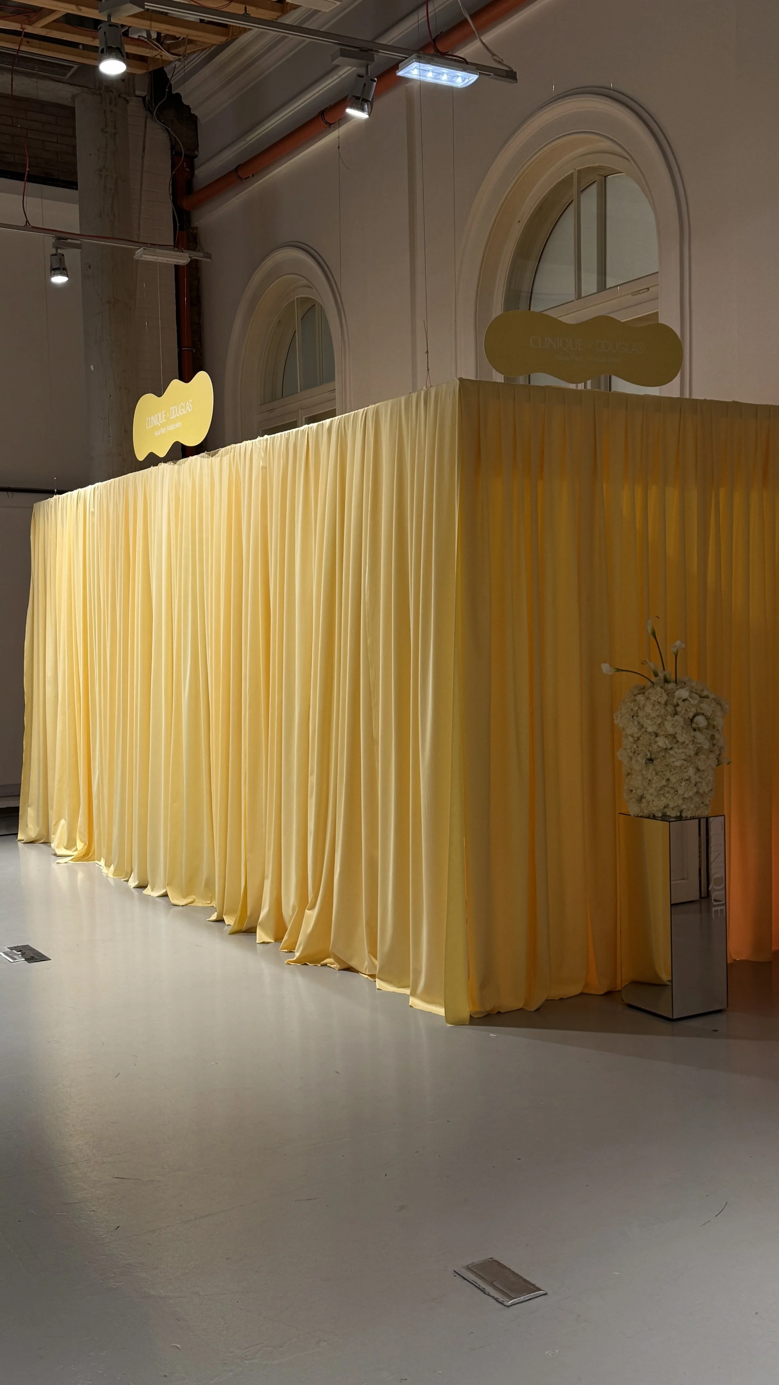 Yellow curtains creating a backdrop at an indoor event space, with a flower arrangement on a small pedestal to the side and signs hanging on the wall above, indicating a Clinique Douglas event.