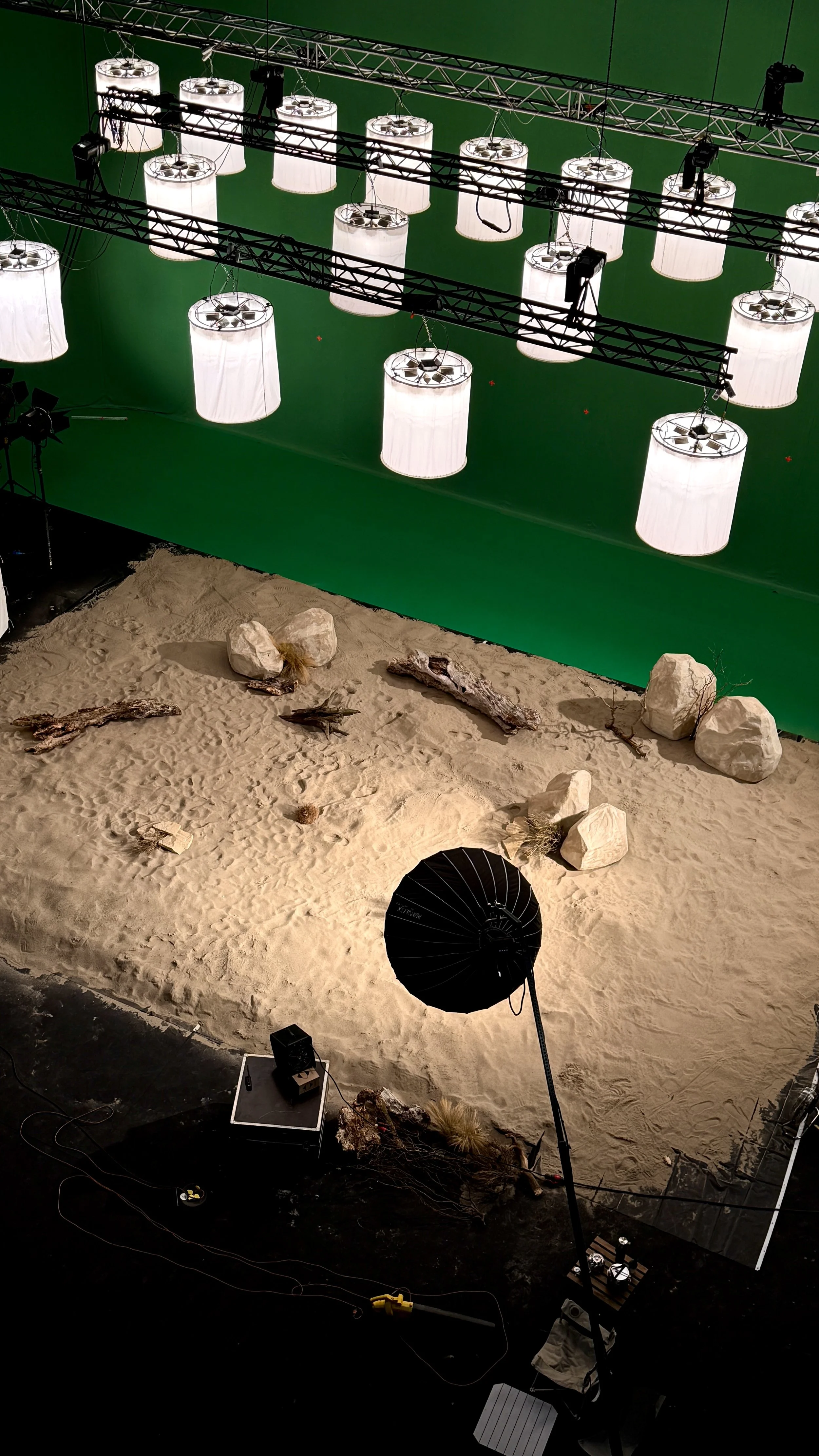 Photo of a photography studio with a green screen background, hanging lamps, studio lights, sand, rocks, driftwood, and a large black umbrella.