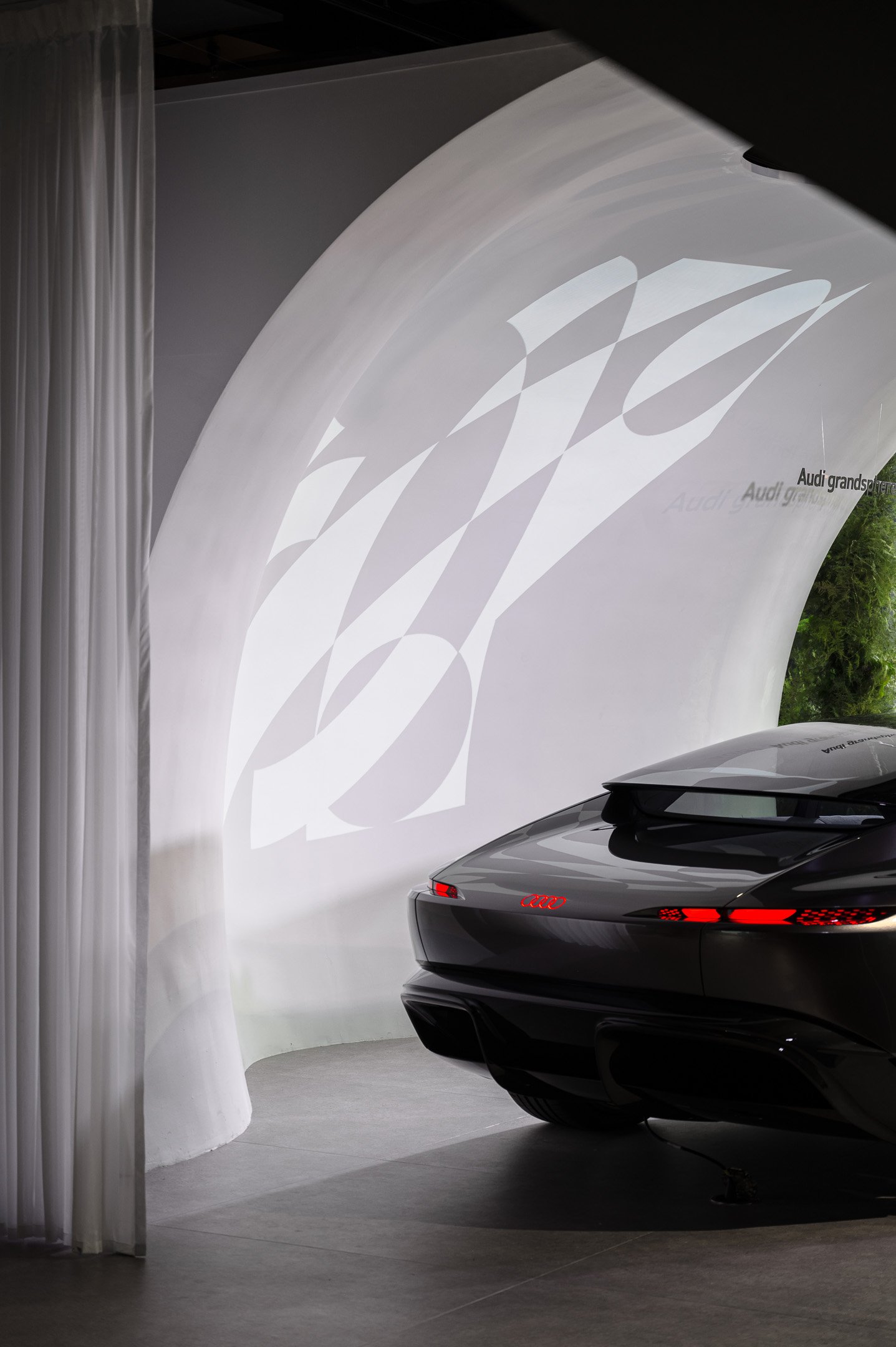 A black sports car with sleek design and red taillights parked indoors near a white curved wall with a large Audi logo projected on it.