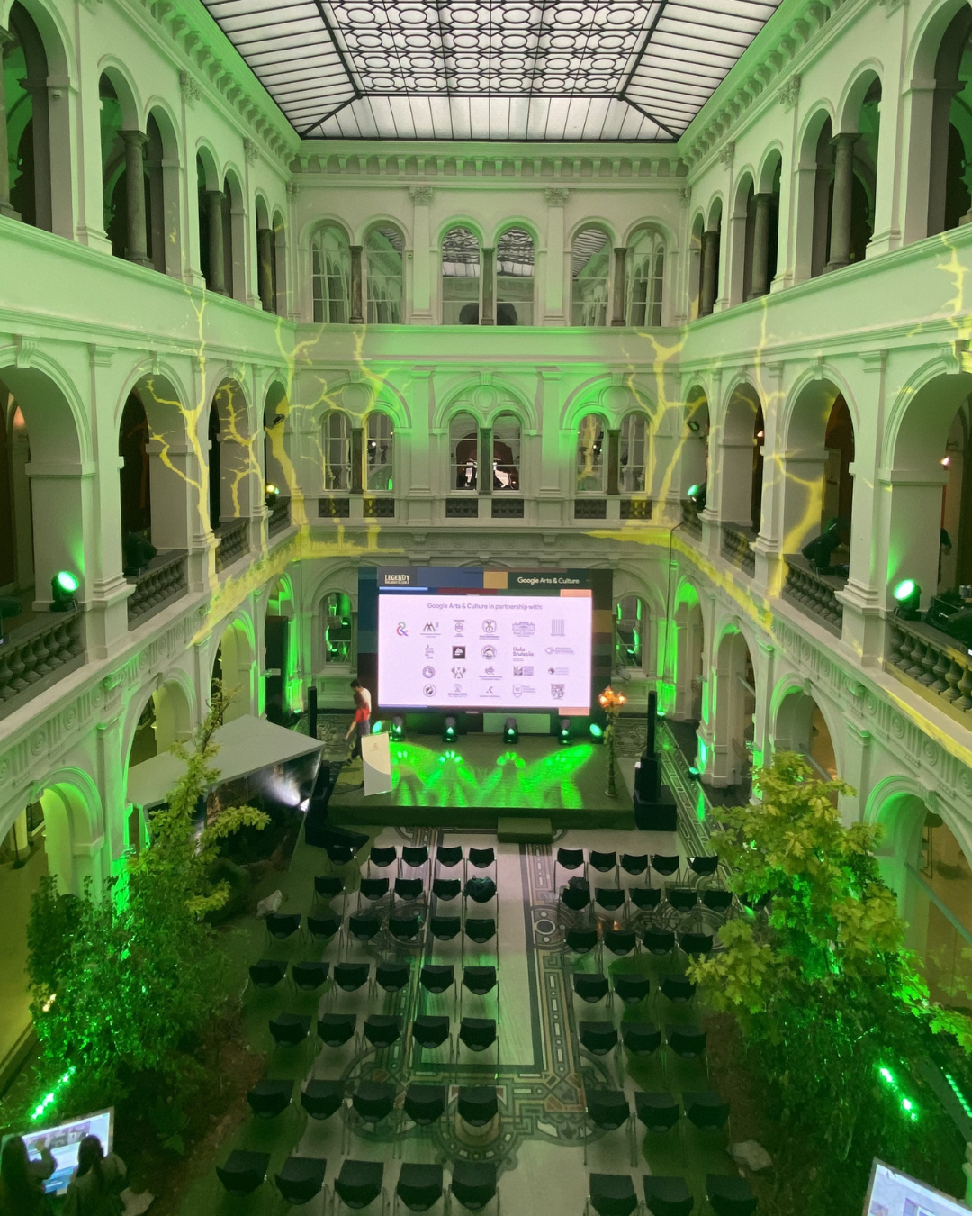 An indoor event space with multiple floors, green lighting, and a large digital screen at the front with chairs arranged in rows.