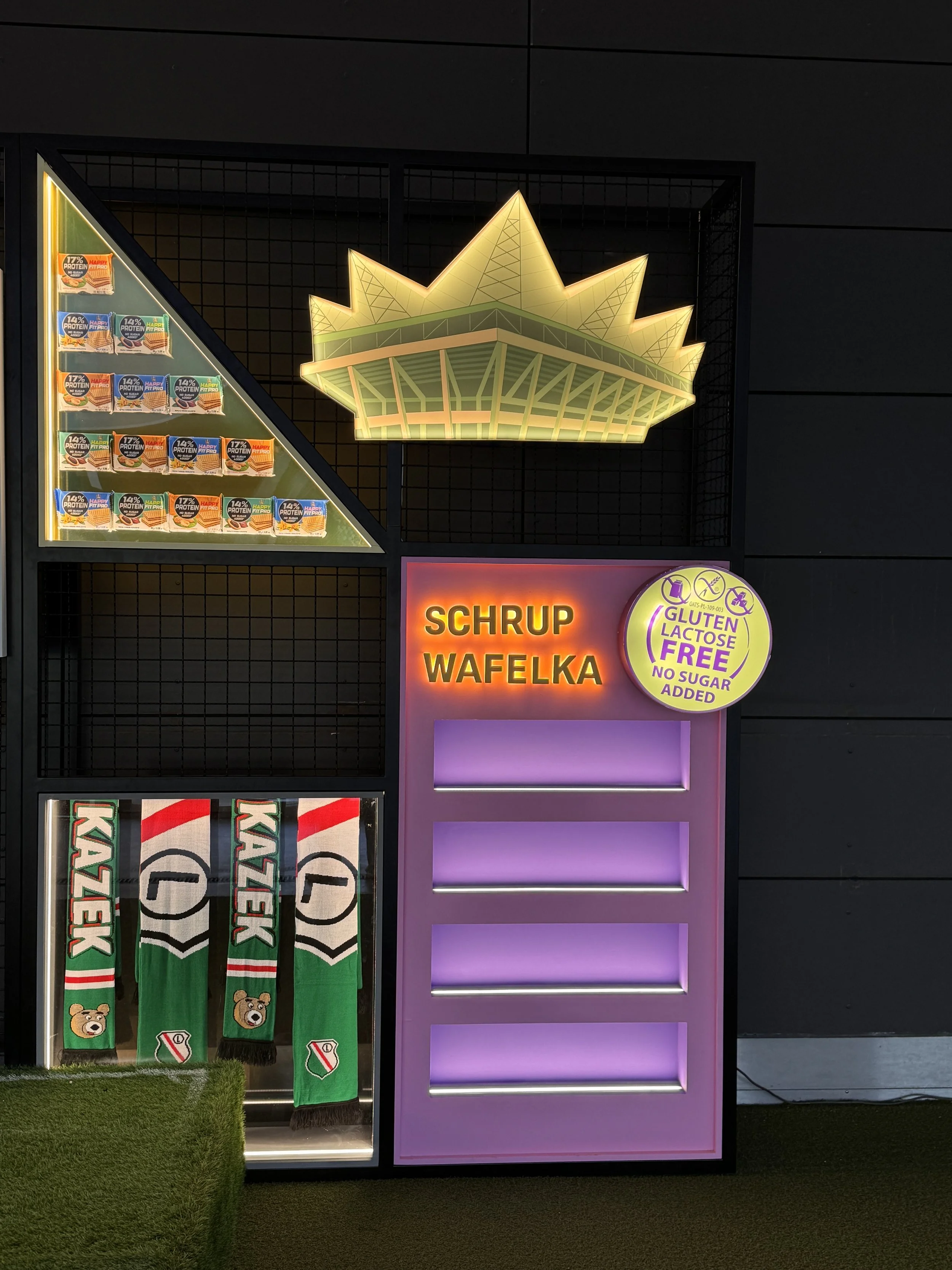 Illuminated neon sign in the shape of a crown, with a menu board below displaying 'Skrup Wafelka' and a yellow circular badge indicating gluten and lactose free, no sugar added. Green stockings with teddy bear and shield designs hanging below.