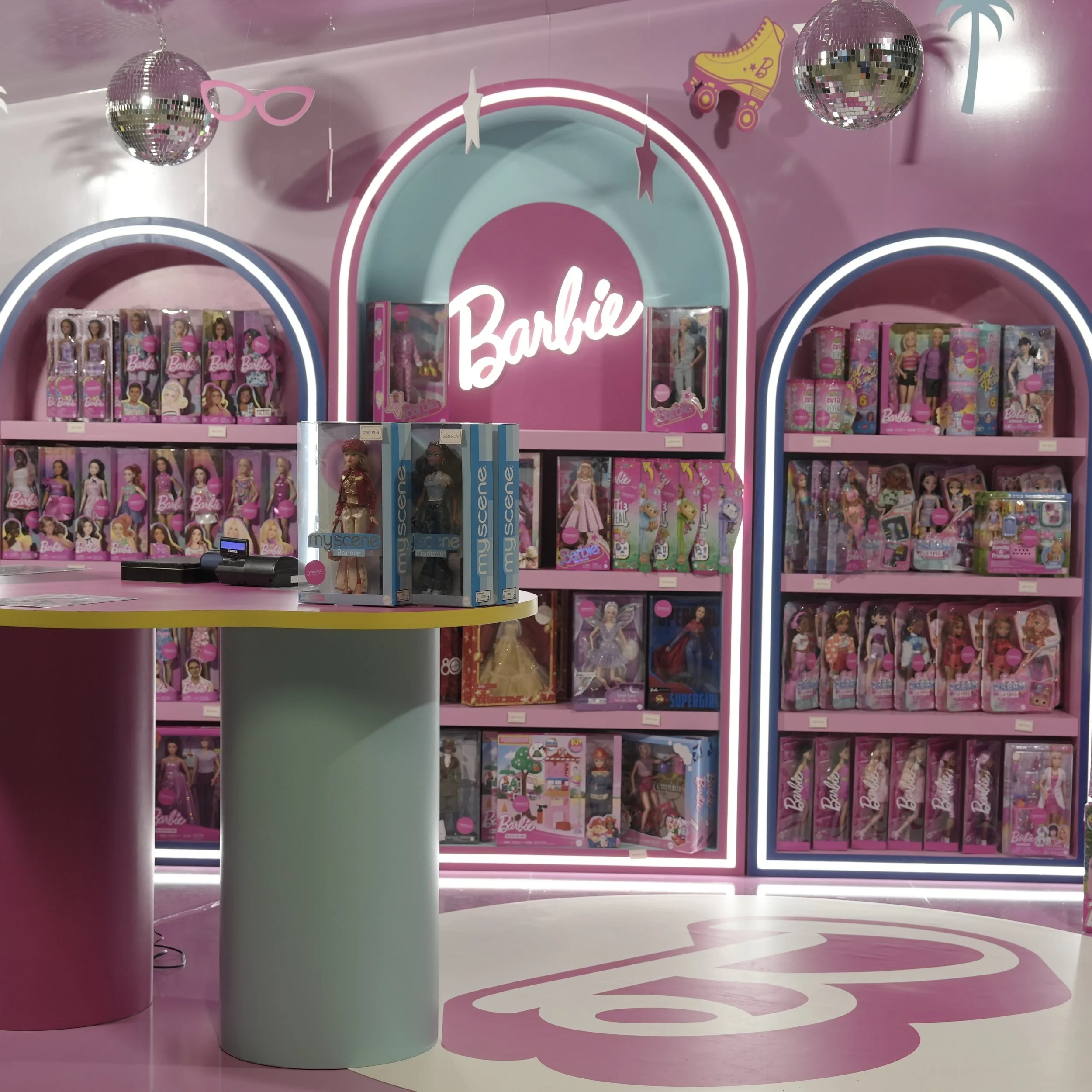 Colorful retail display with Barbie dolls in pink and purple boxes on pink shelves, neon Barbie sign, disco ball decorations, and a pink roller skate graphic in the background.