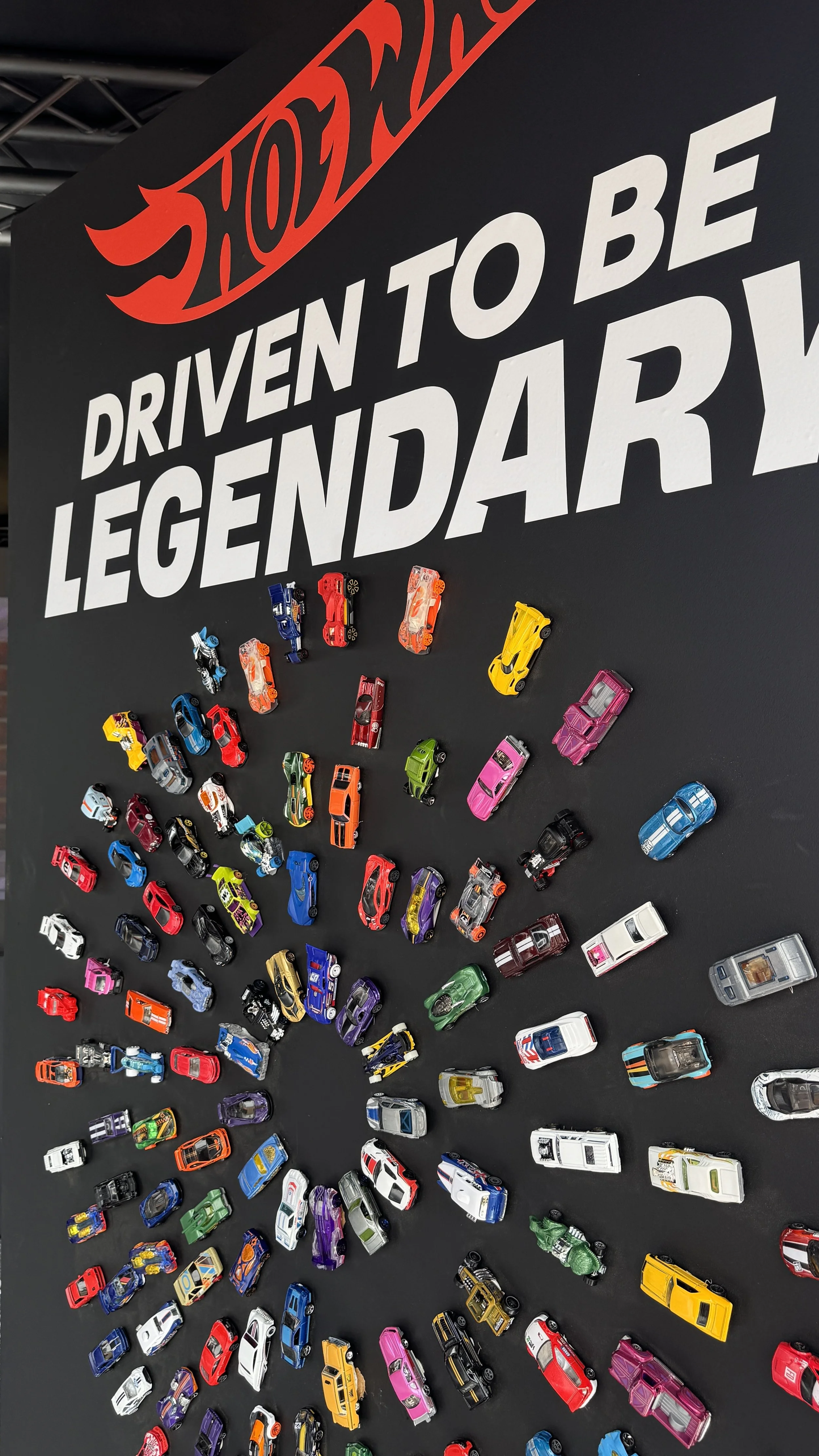 Display of various toy cars arranged on a black surface with the Hot Wheels logo and the phrase 'Driven to be a legend' on a sign background.