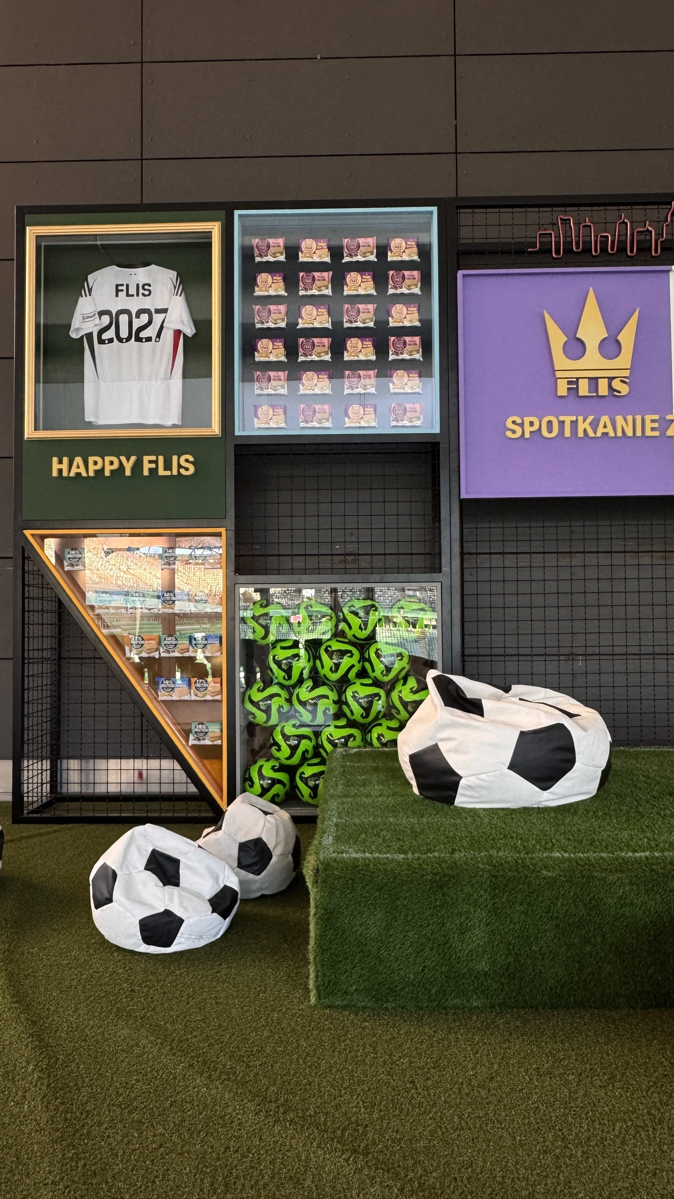 Display with a framed soccer jersey reading 'FLIS 2027', motivational signs, soccer equipment including multiple green and black soccer balls, and soccer-themed bean bags on artificial turf.