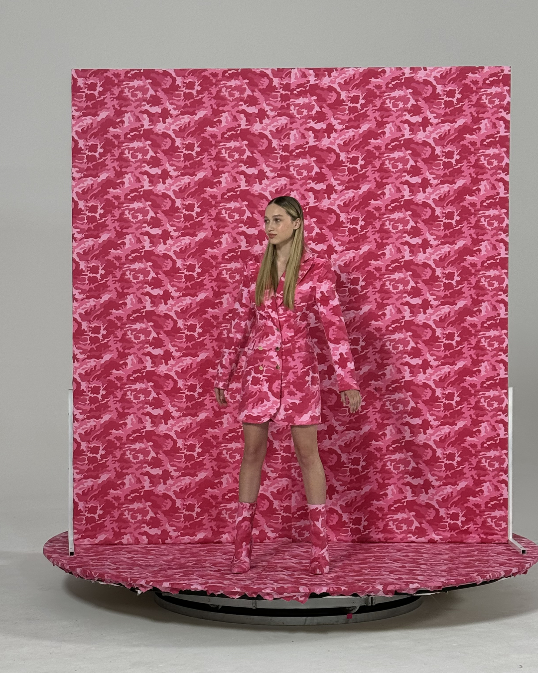A woman stands on a circular platform in front of a pink camouflage-patterned backdrop, wearing a matching pink camouflage coat, shorts, and boots.