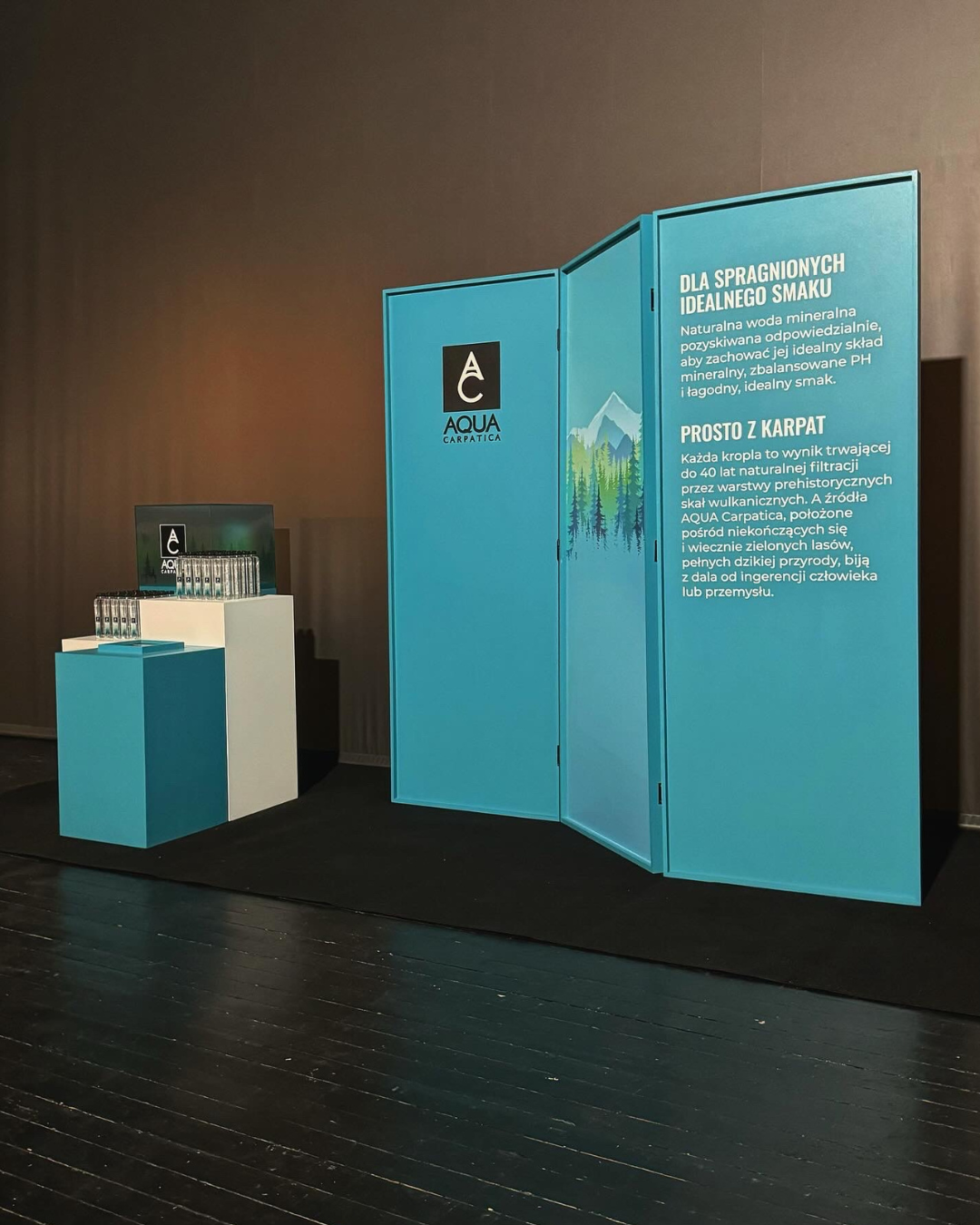 Indoor display with a blue folding informational panel and a small table with bottled water and branded materials. The panel has the AQUA Carpatica logo and text in Polish about mineral water sourced from the Carpathians.