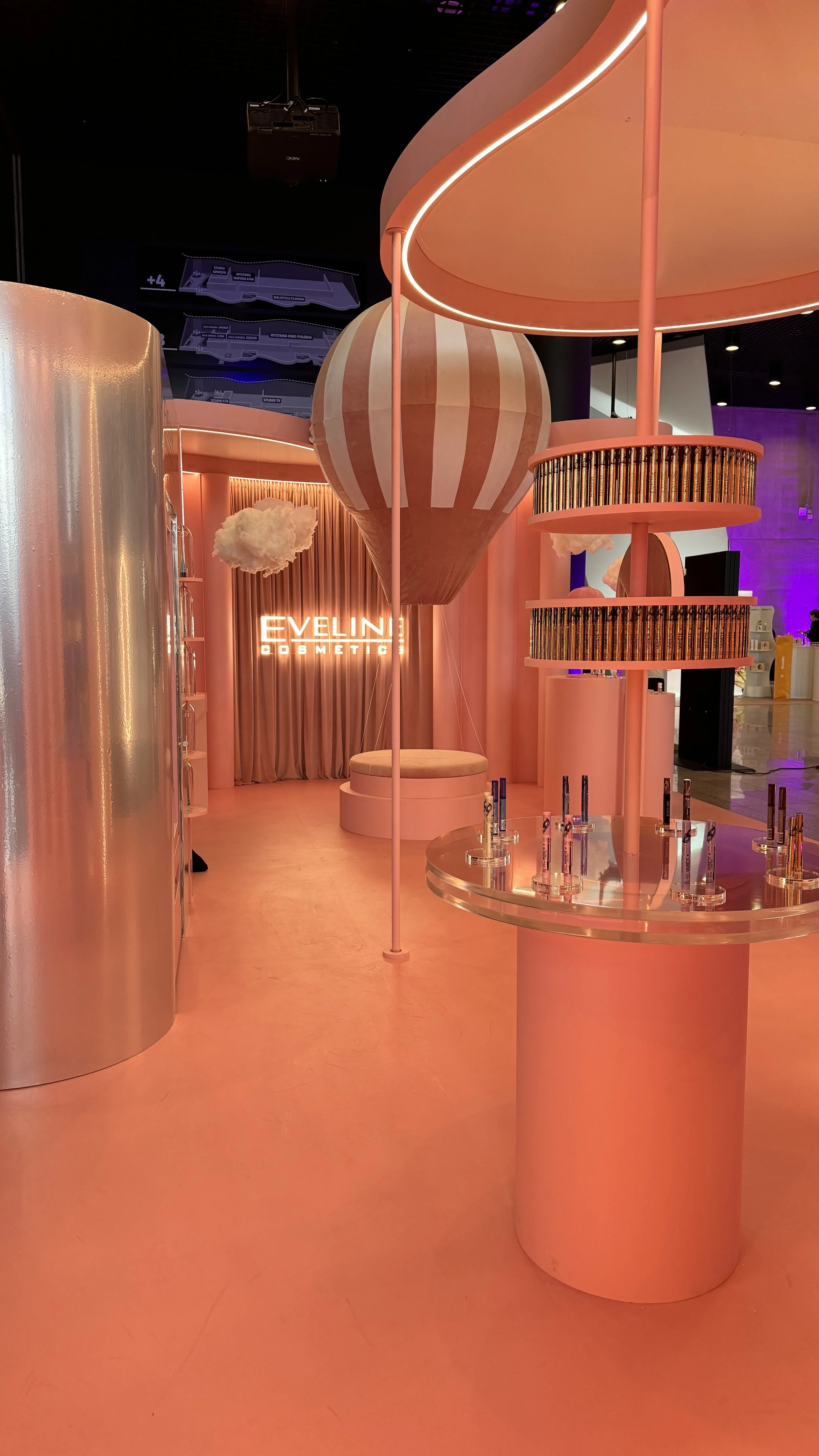 Interior of a cosmetic store with pink and peach decor, display counter with beauty products, illuminated sign reading 'EVELINE COSMETIC,' and decorative clouds hanging from the ceiling.