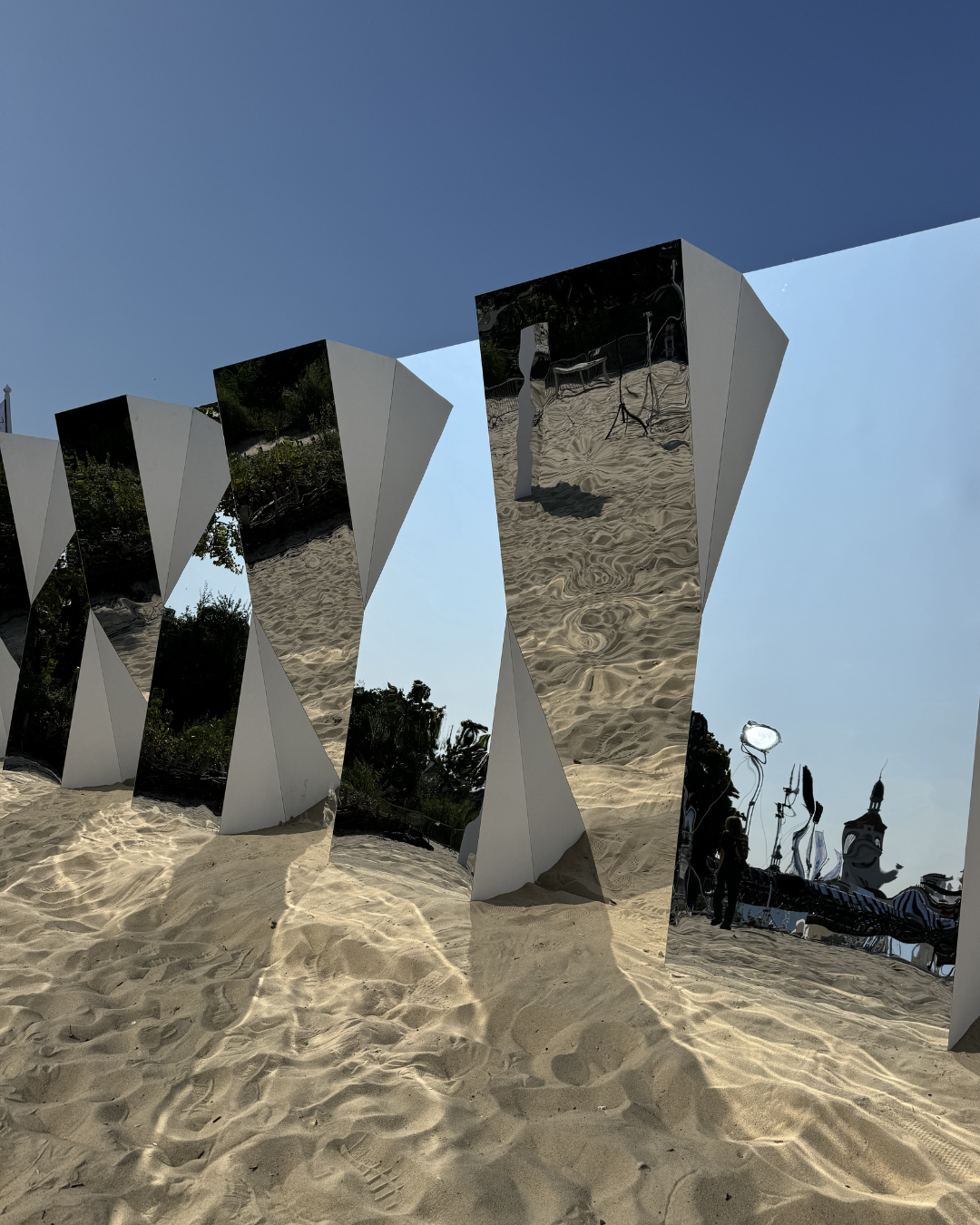 Reflective metal sculptures on sandy ground with a clear blue sky in the background