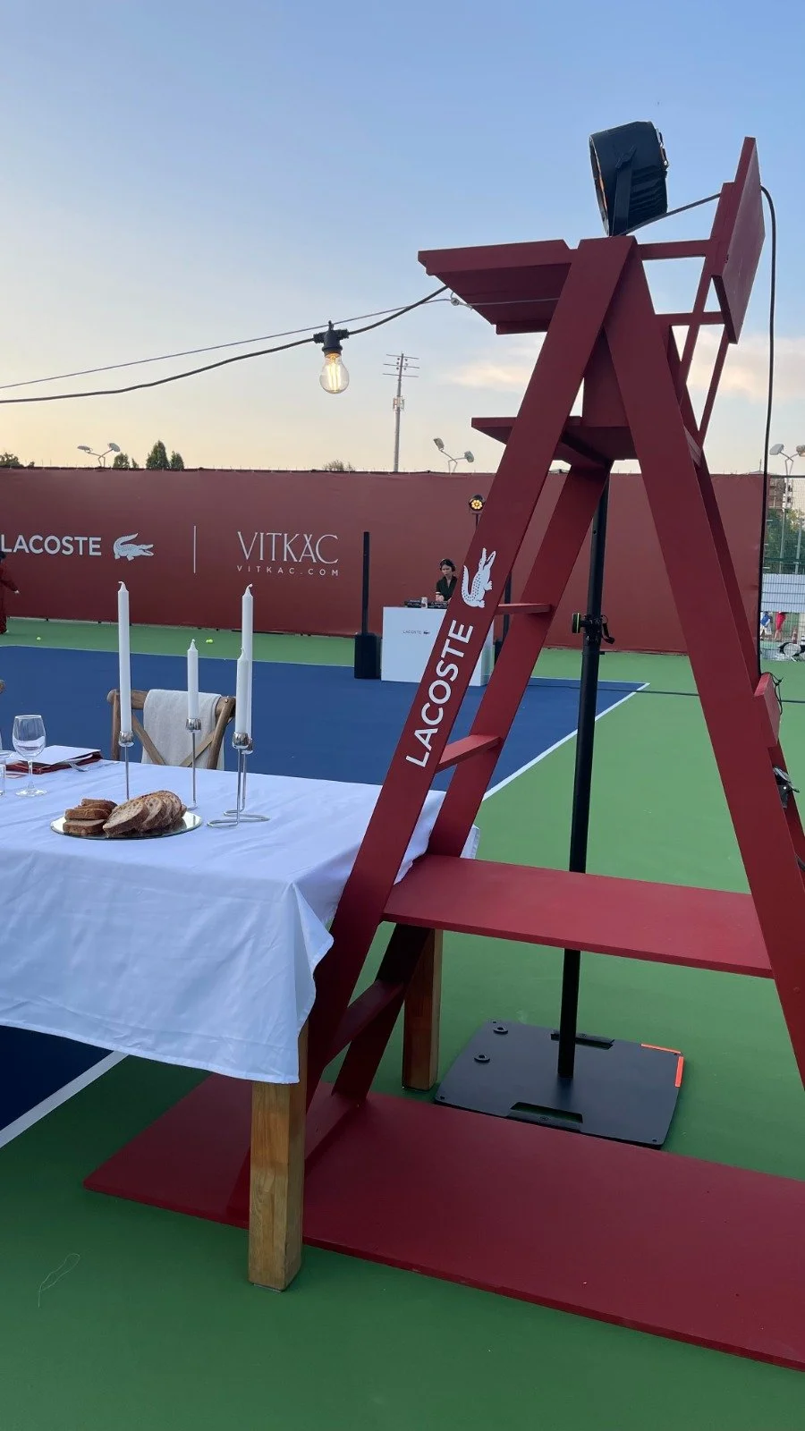 A tennis court with a red Lacoste sign, a table set for dining with candles, bread, wine glasses, and a red ladder structure with Lacoste signage and a crocodile logo, under a clear sky.