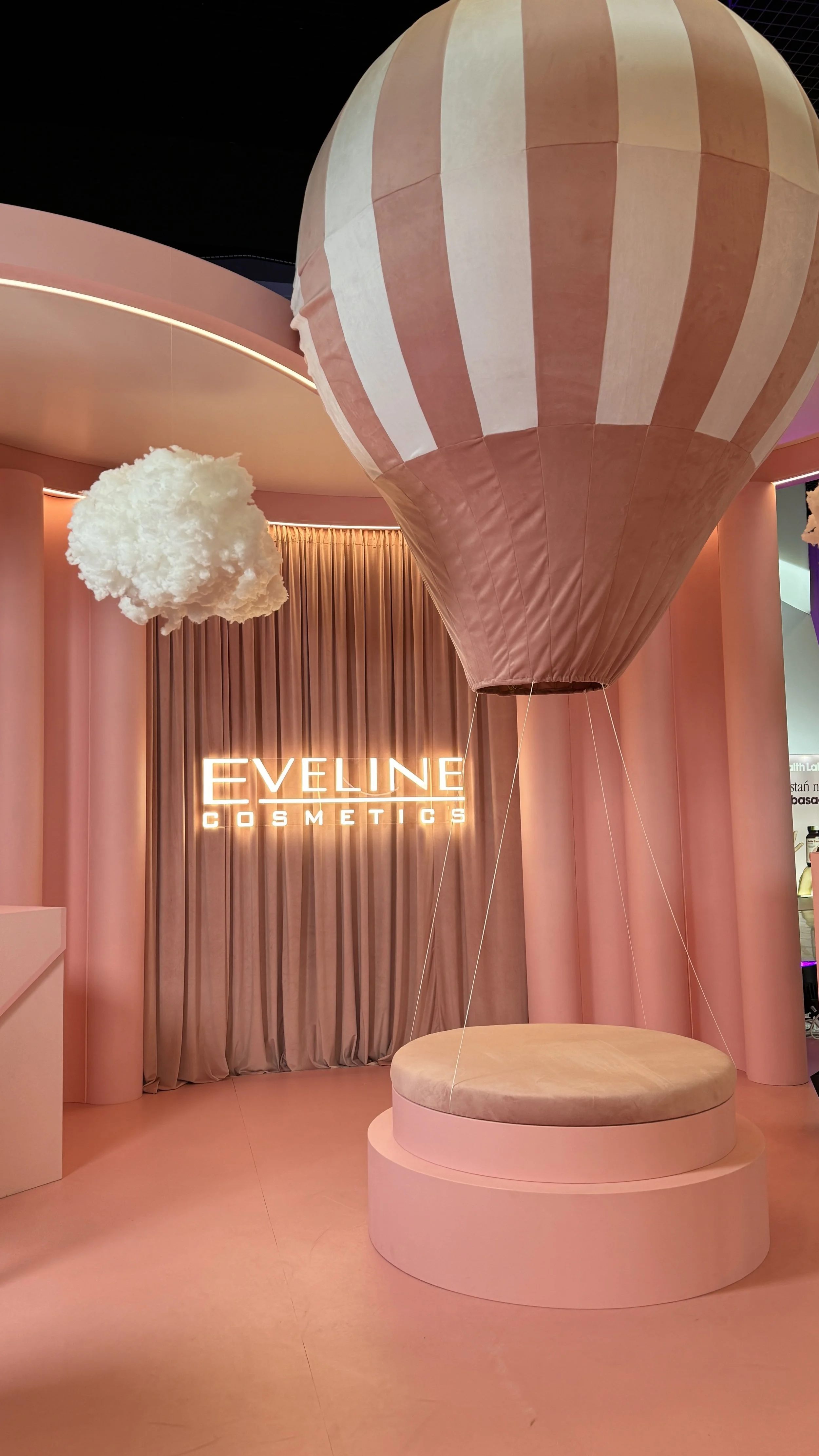 Inside a pink-themed store with a hot air balloon decoration, a fluffy white cloud decoration, and illuminated "EVELINE COSMETICS" sign.