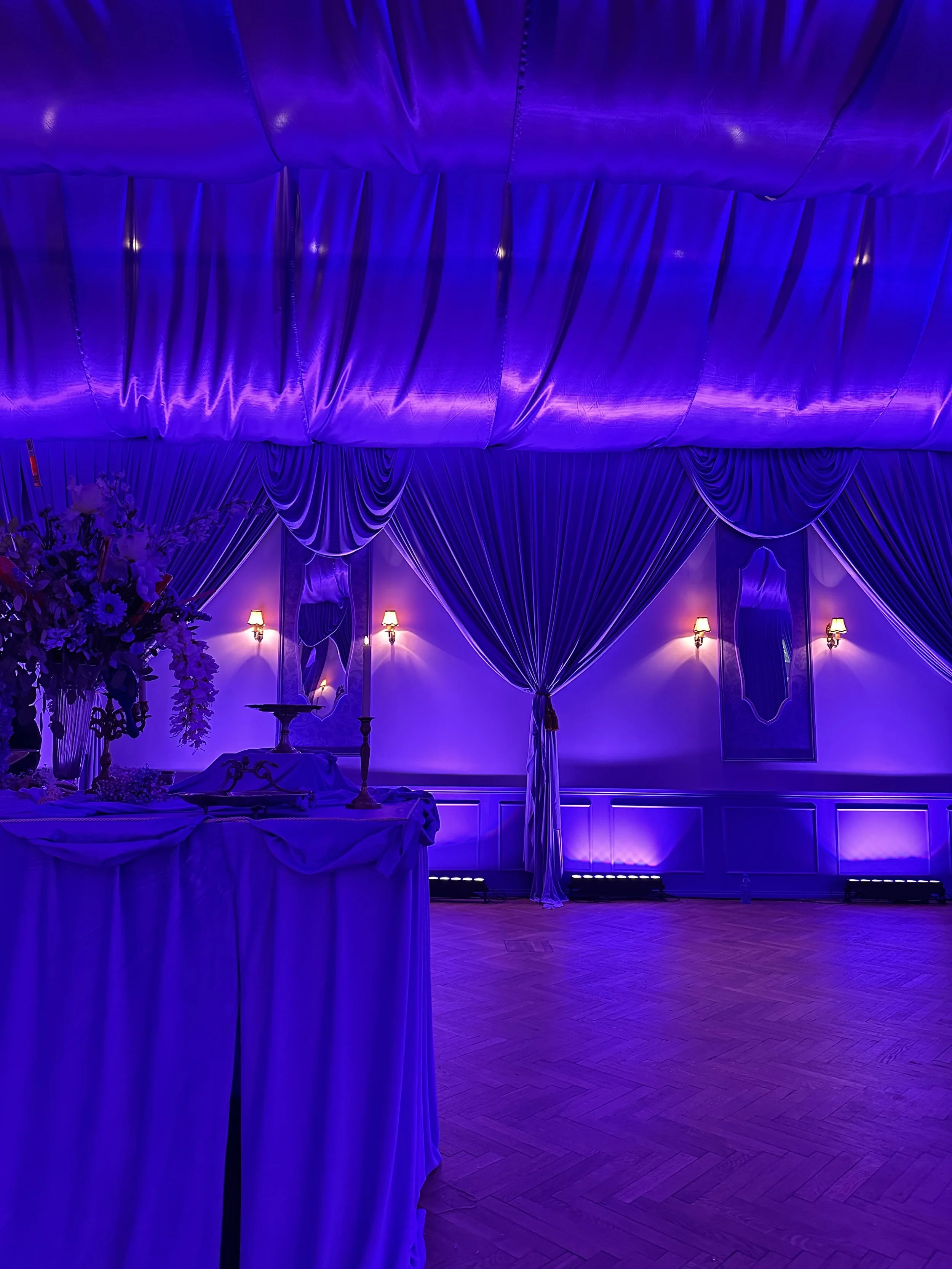Elegant banquet hall decorated with purple lighting, draped curtains, and a table with floral centerpieces and candles.