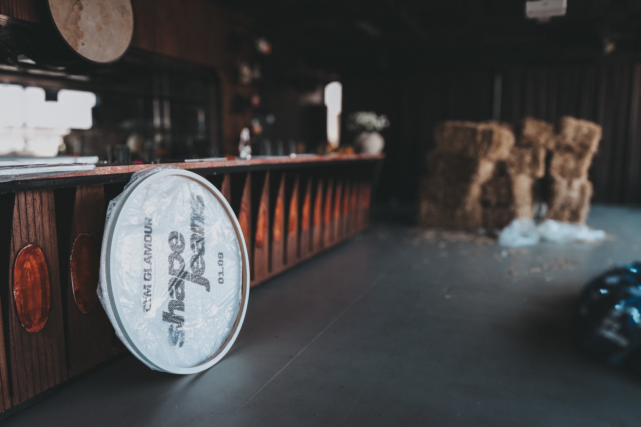 Close-up of a round plastic disc with text, leaning against a wooden railing in a rustic setting. In the background, straw bales are stacked in a barn-like structure. The scene appears to be in a farm or rural environment.
