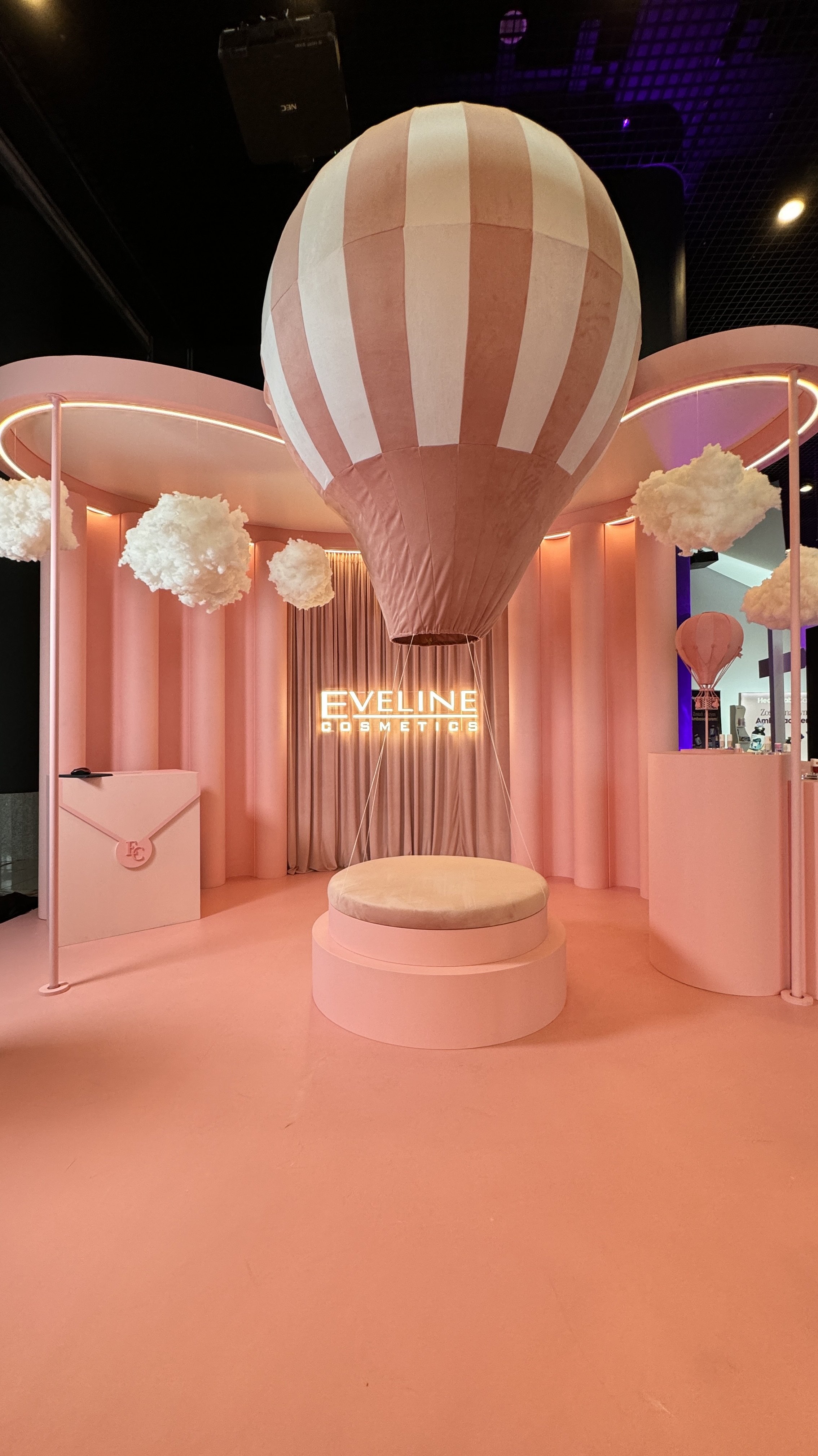 Interior of a pink-themed booth for Eveline Cosmetics with a hot air balloon decoration, cloud props, and a neon sign displaying the brand name.