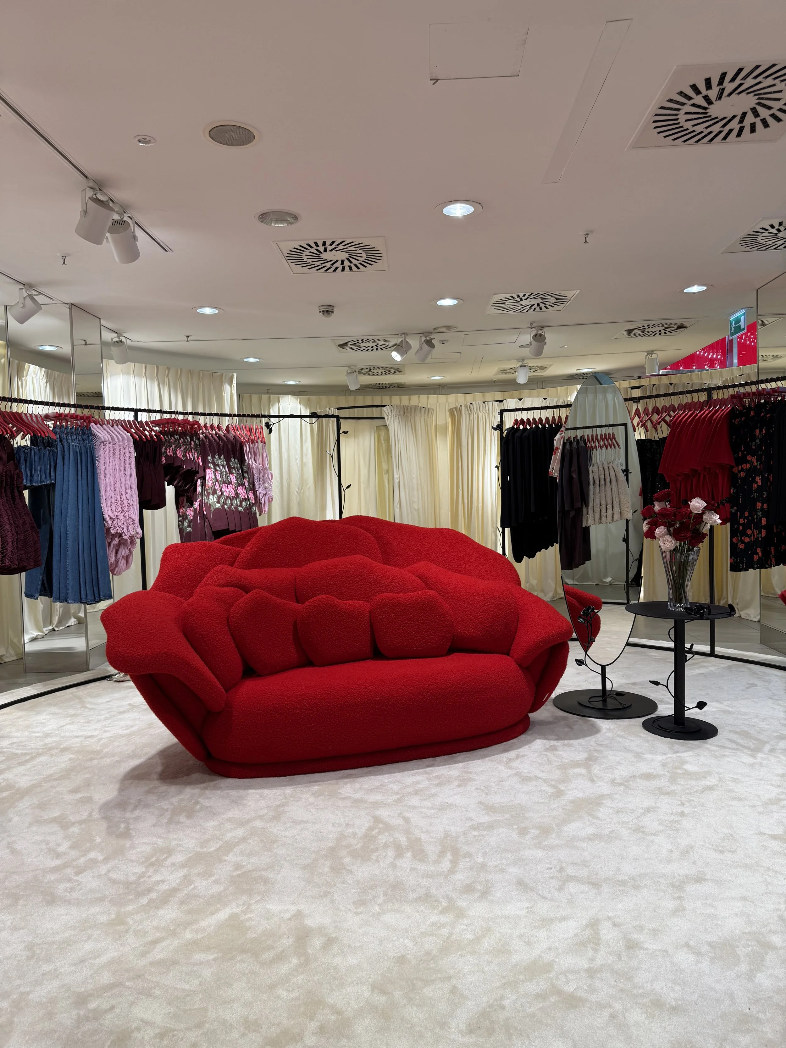 A red plush sofa situated in a clothing store fitting room surrounded by racks of dresses, with a mirror to the right and decorative flowers on a small table beside it. The ceiling has various lights and ventilation panels.