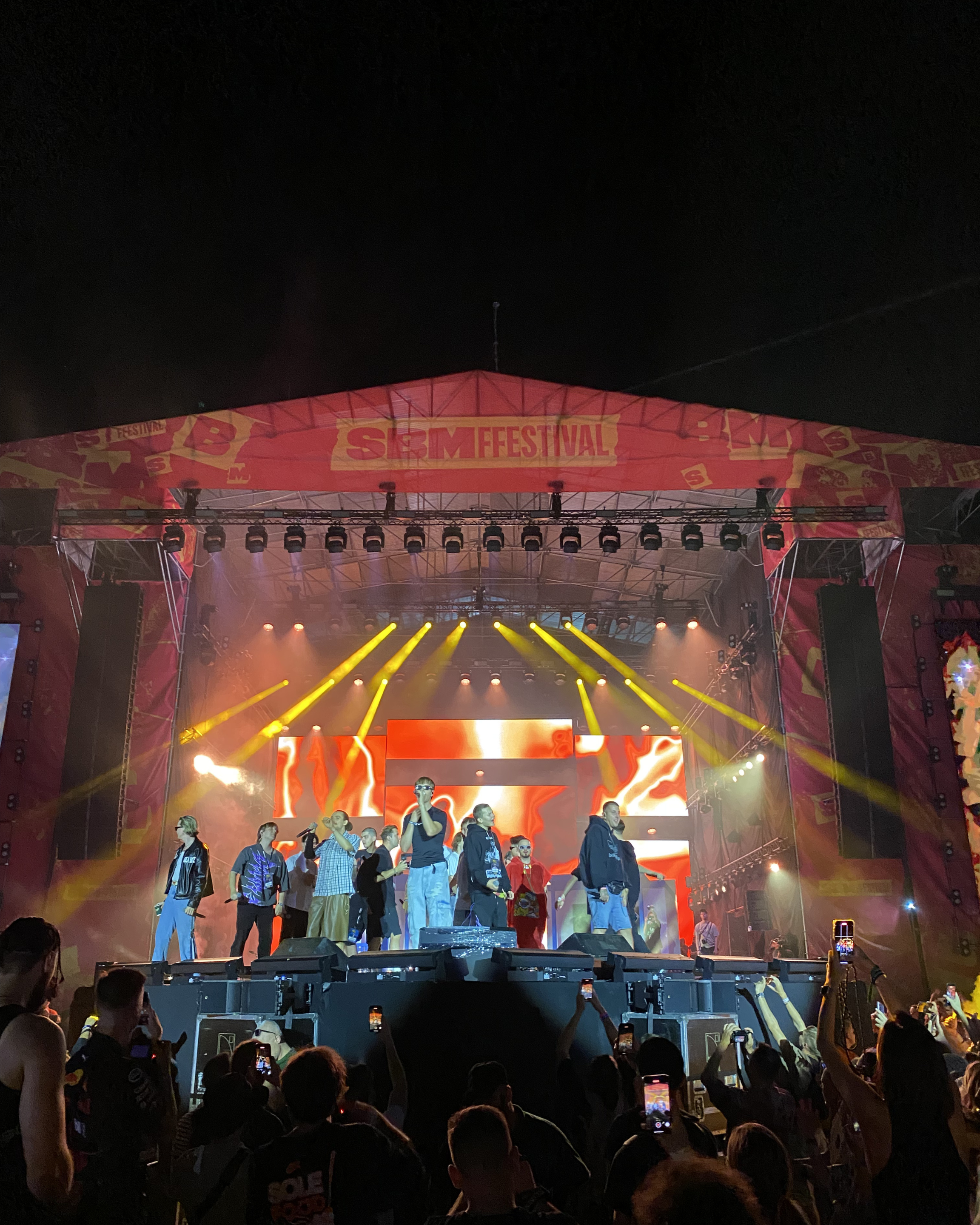 A live music concert on a large outdoor stage at night, with performers and performers, stage lights, and a crowd of audience members taking photos and videos.