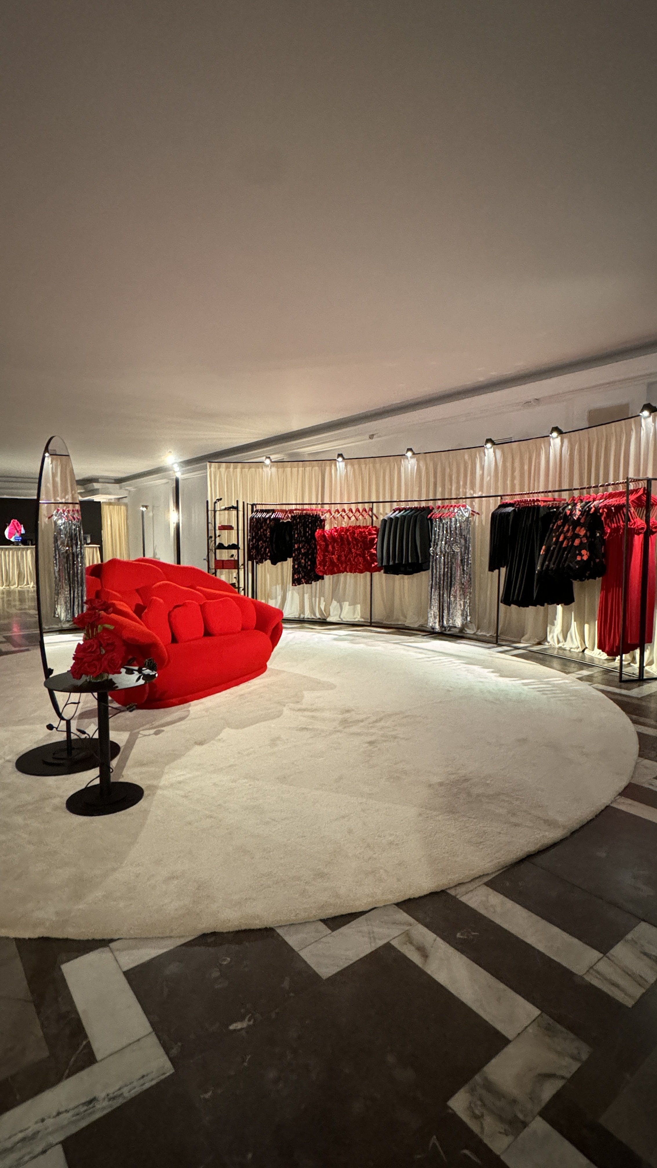 A boutique with a red sofa, a small black side table, and clothing racks displaying various dresses and garments against a cream-colored curtain backdrop.