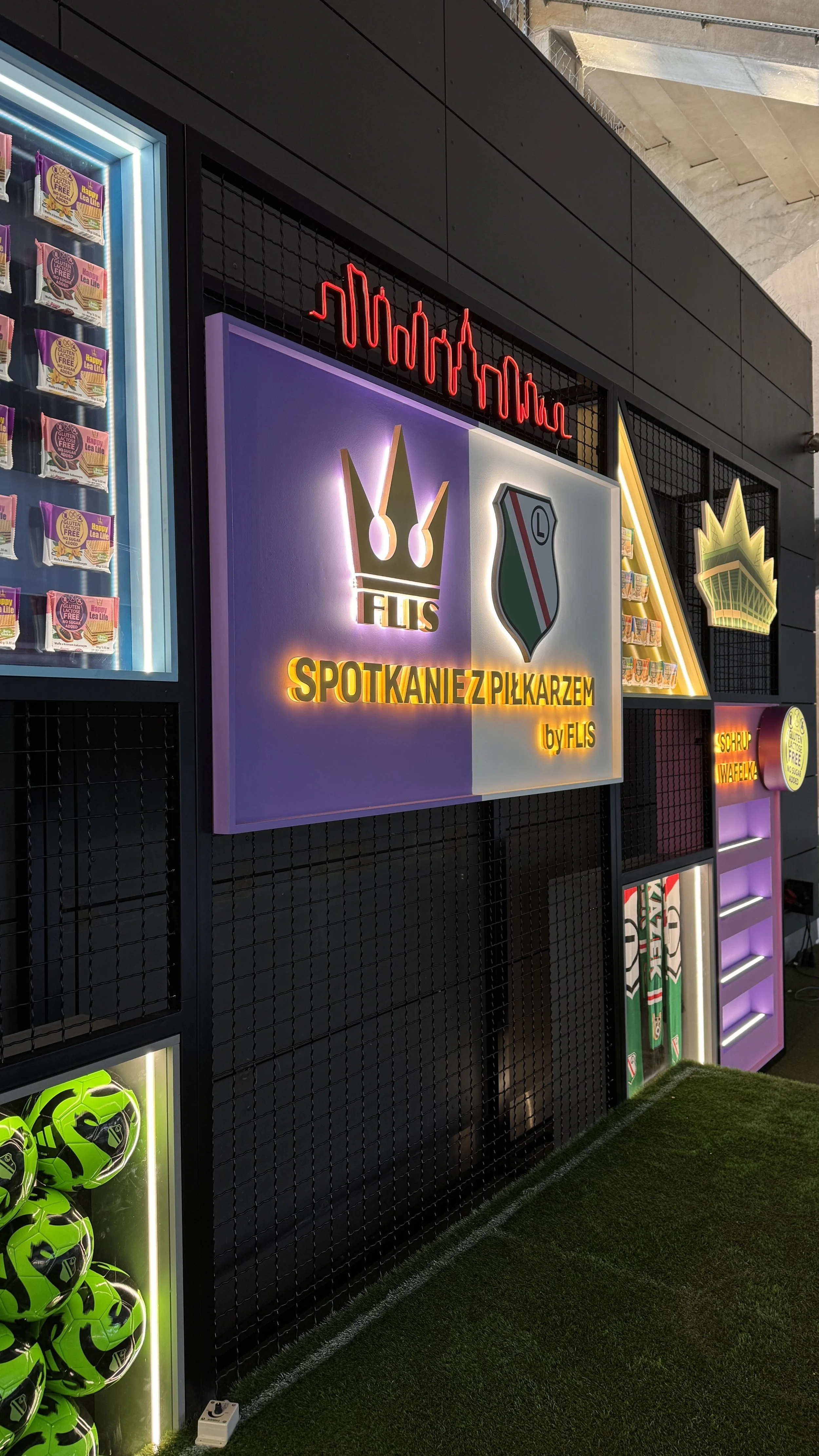 A display wall with illuminated signs and sports memorabilia, including colorful signage in Polish about soccer players, and a collection of green soccer balls.